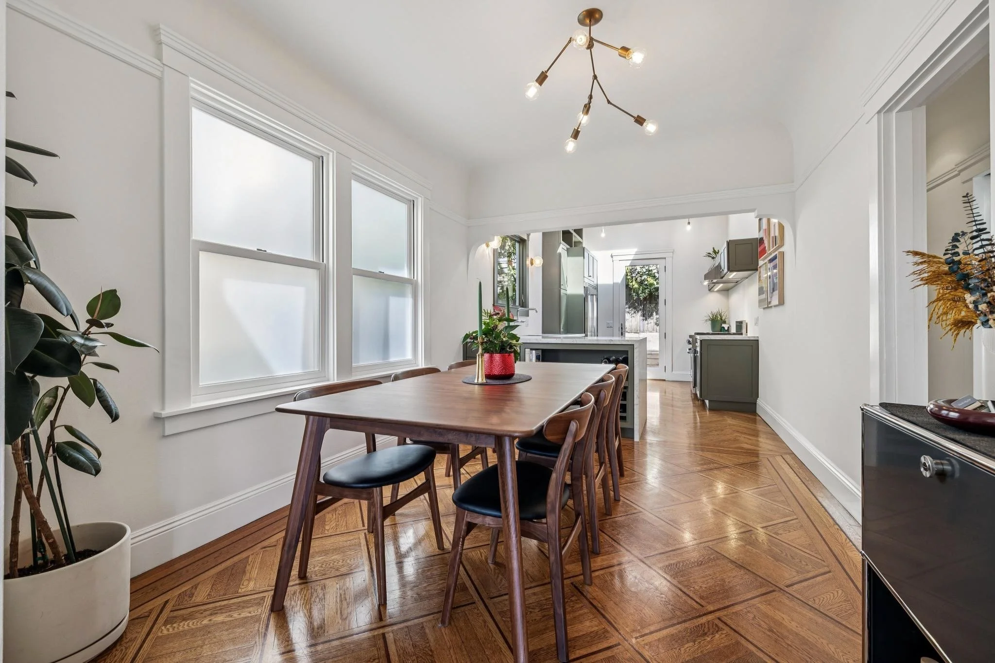 Dining room with wooden table and chairs, large windows, hardwood floor, modern chandelier, and view into kitchen area with gray cabinets and door to backyard.