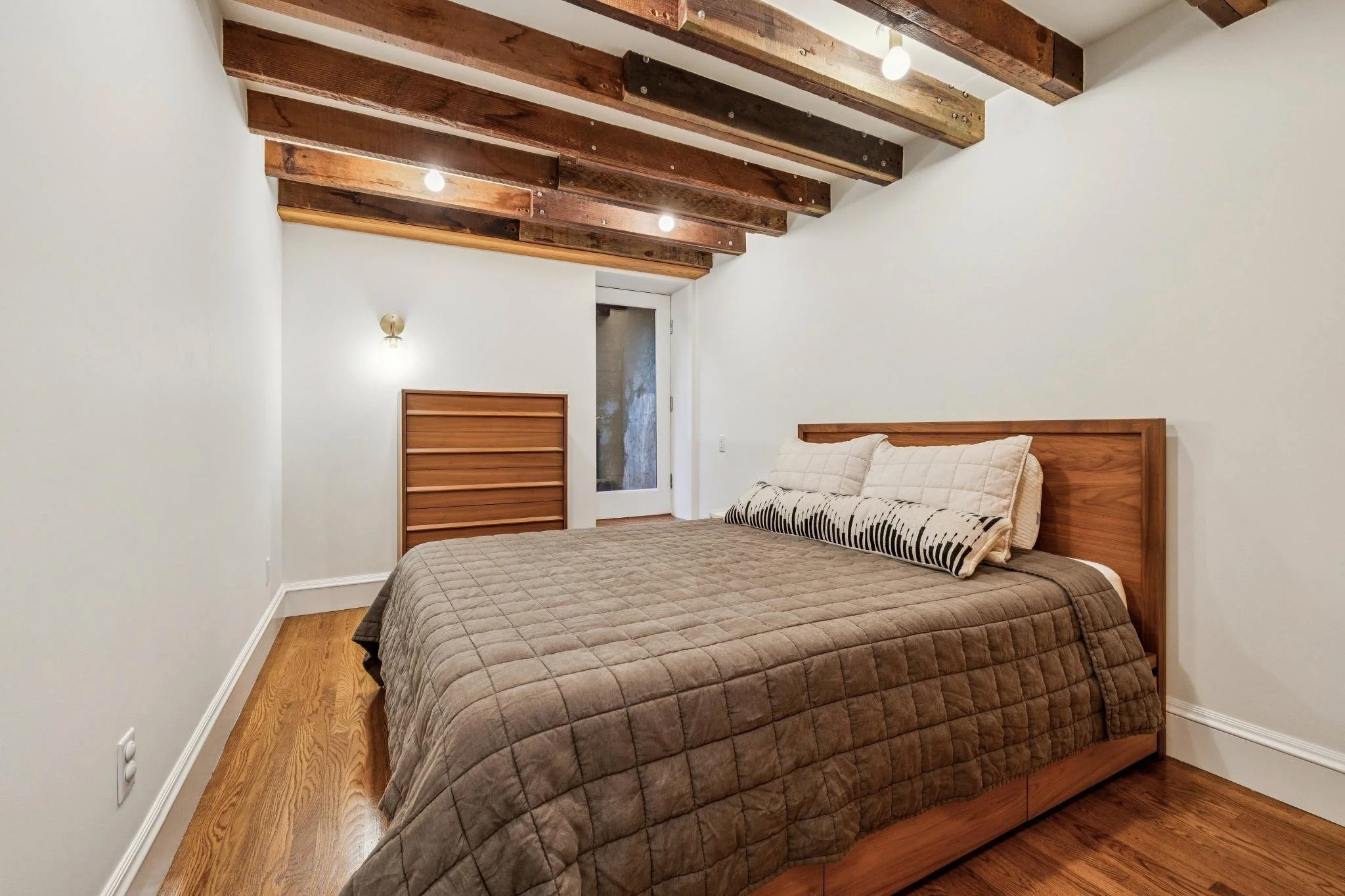 A bedroom with a wooden bed frame a beige quilted bedspread and pillows, wooden ceiling beams, a wall sconce, and glass door leading outside.