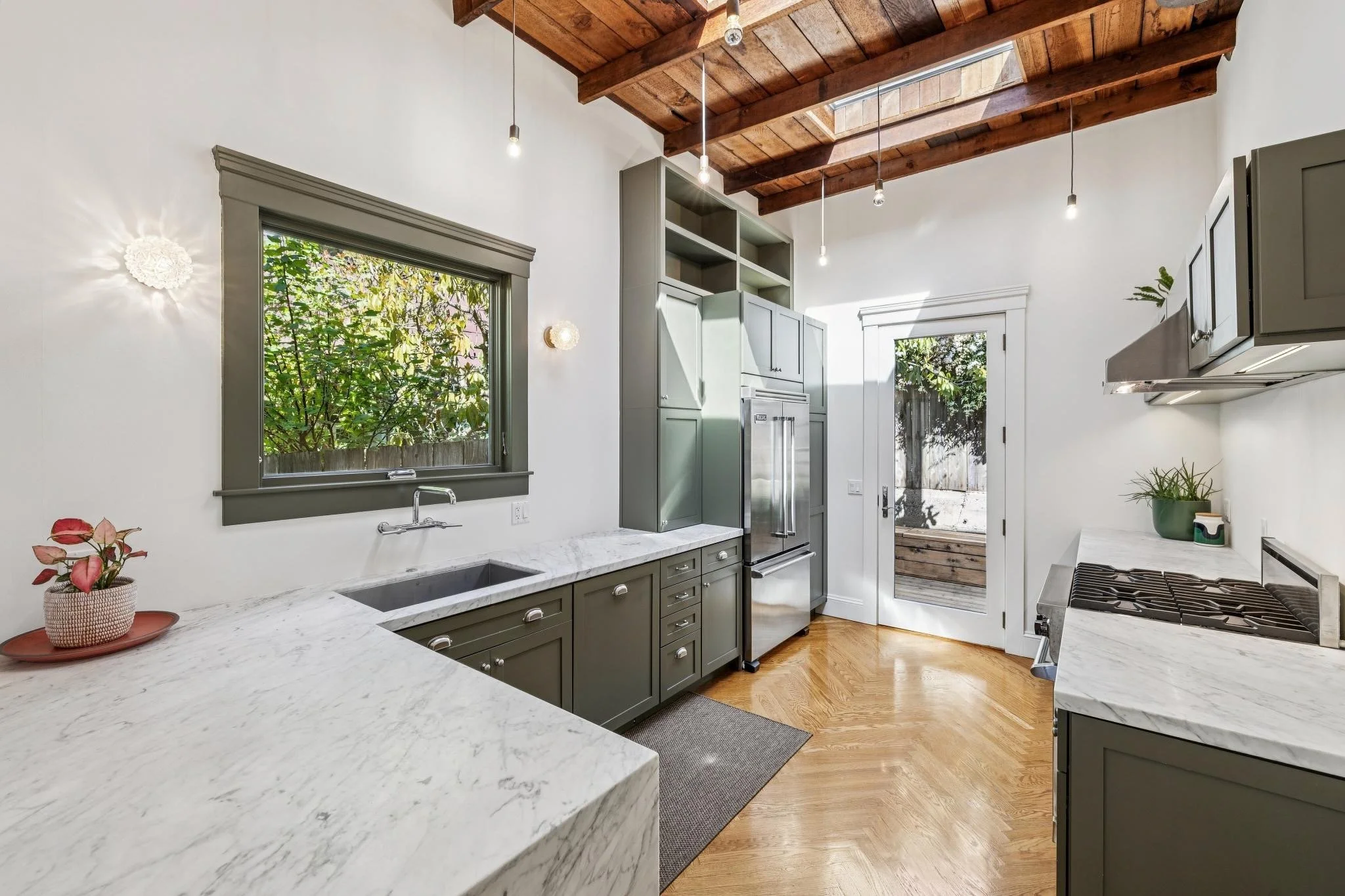 Modern kitchen with grey cabinetry, white marble countertops, stainless steel refrigerator, stove, and wood ceiling with skylights, surrounded by white walls and hardwood floor.