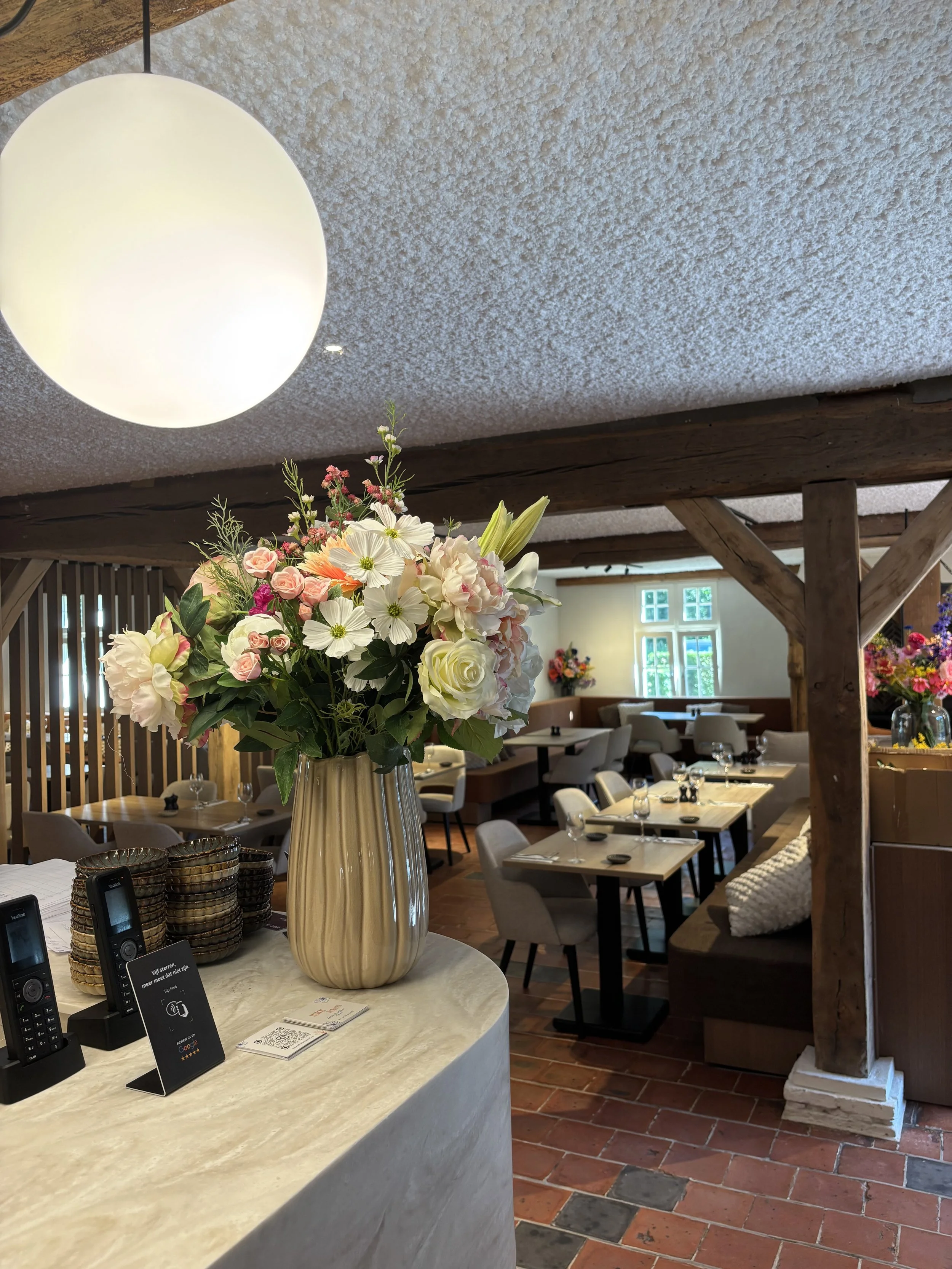Interior of a restaurant or cafe with a large floral arrangement in a beige vase on a white marble counter, and dining tables with chairs in the background. The ceiling has a textured finish, and there is wooden structural support visible.