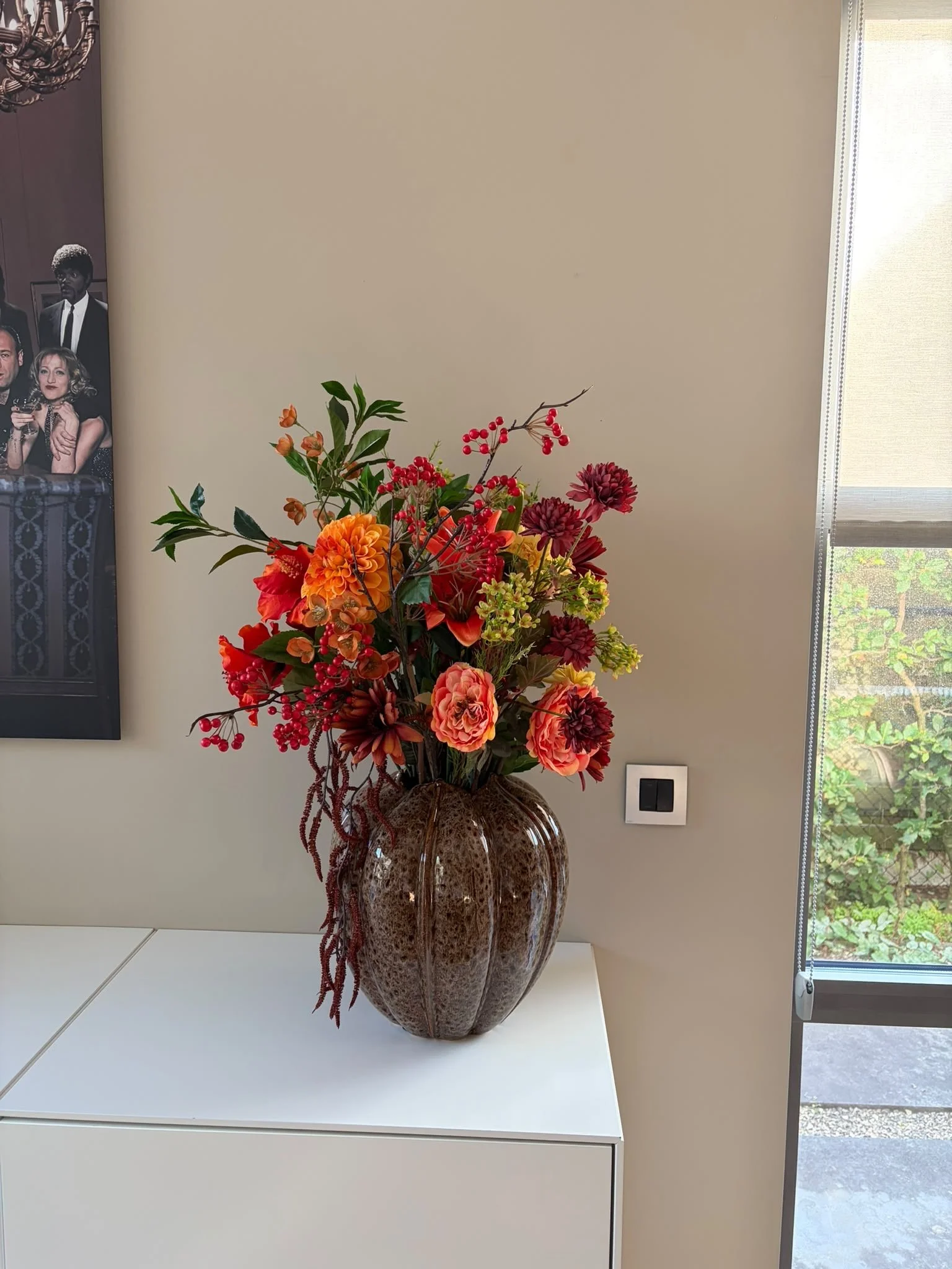 A large, decorative ceramic vase filled with an arrangement of artificial flowers, including orange and red dahlias, red berries, and green leaves, placed on a white table in front of a beige wall and a window with a partly drawn roller blind.