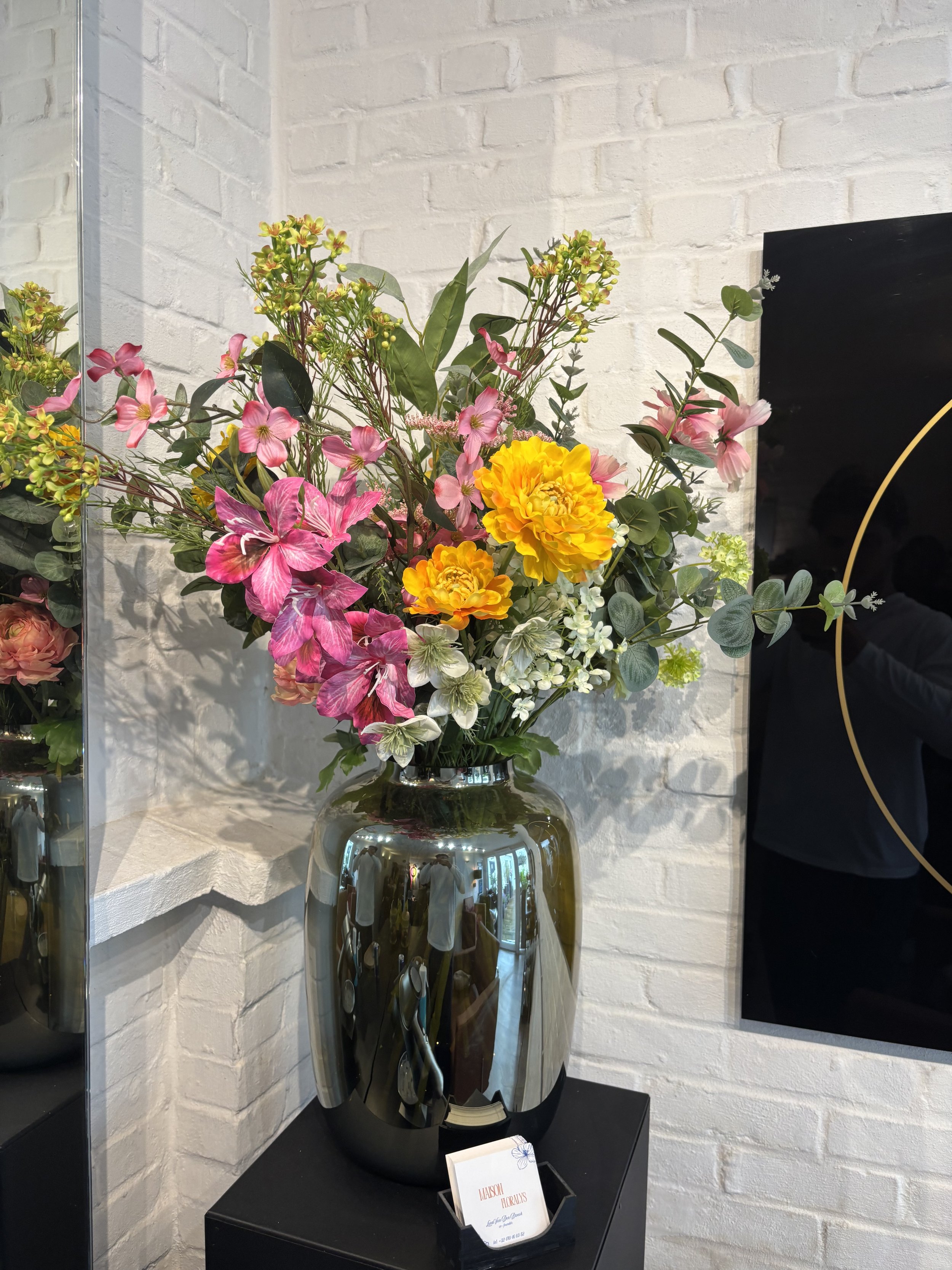 A floral arrangement with yellow, pink, and white flowers in a reflective metallic vase on a black table against a white brick wall.