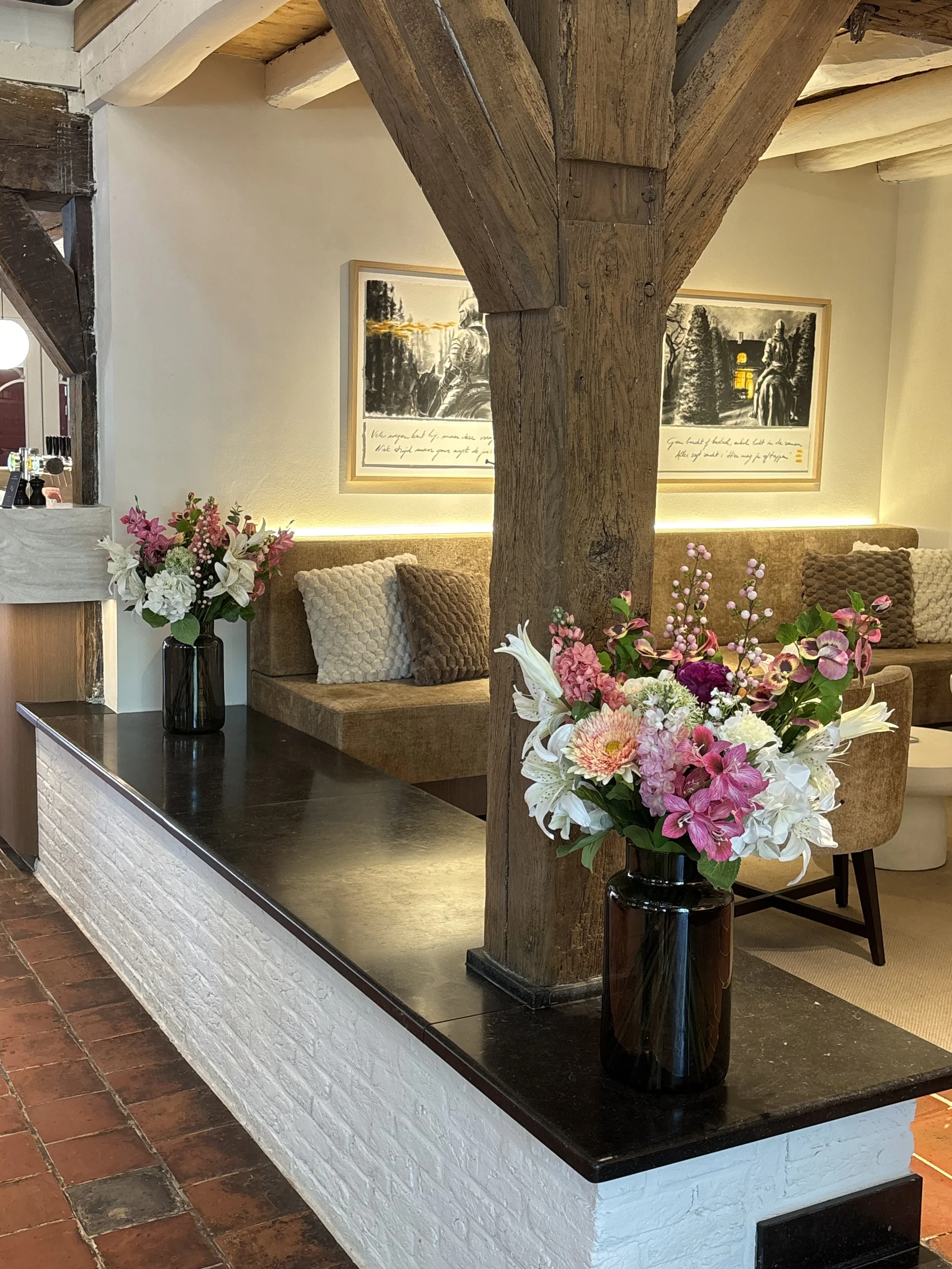 Interior view of a cozy room with a large wooden support beam, a floral arrangement with pink and white flowers in a black vase, framed artwork on the wall, and cushioned seating.
