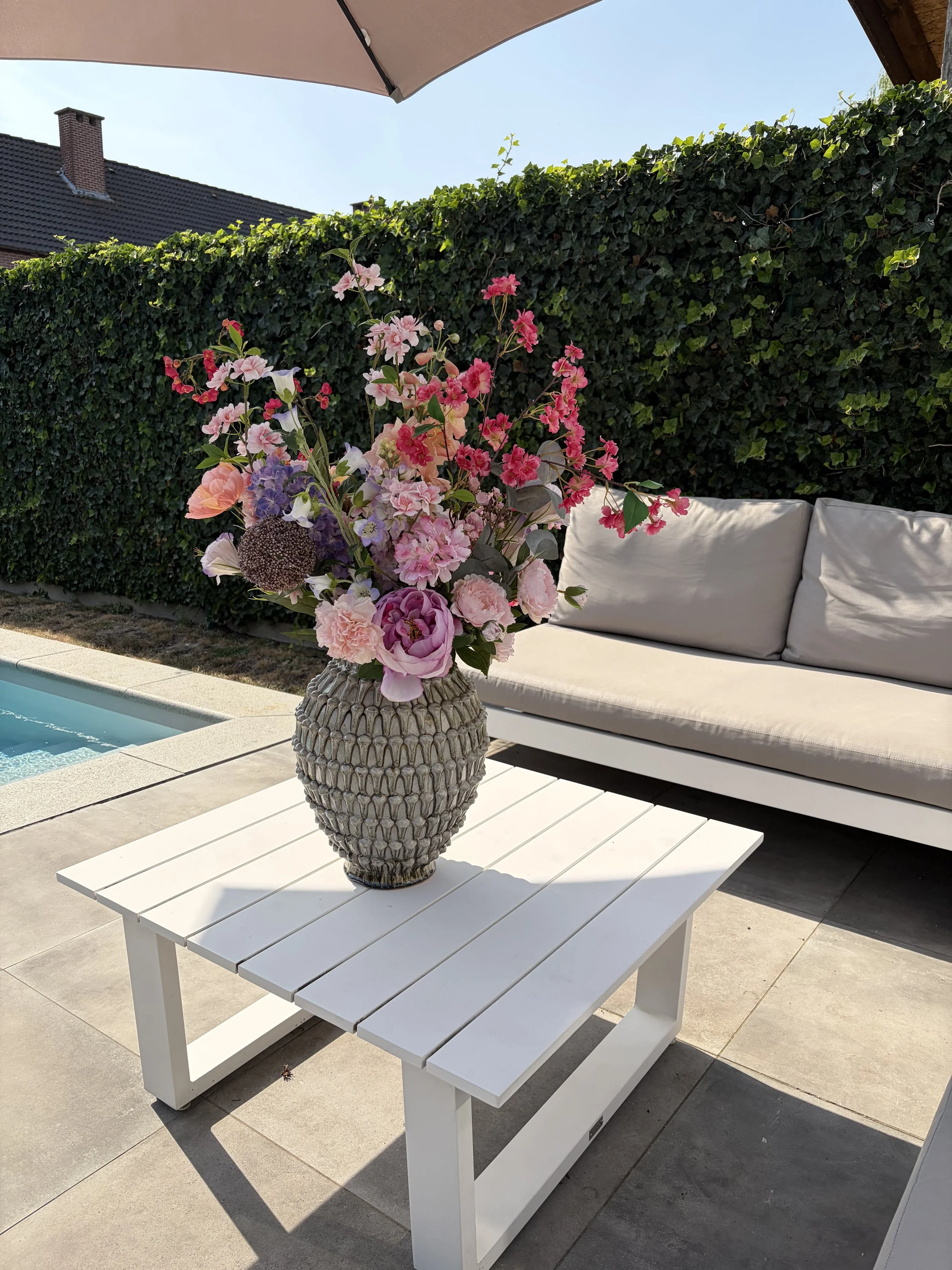 A white outdoor table with a large floral arrangement in a textured gray vase. Behind the table is a white outdoor sofa with cushions, a dark green hedge, a blue sky, and a roof with a chimney.