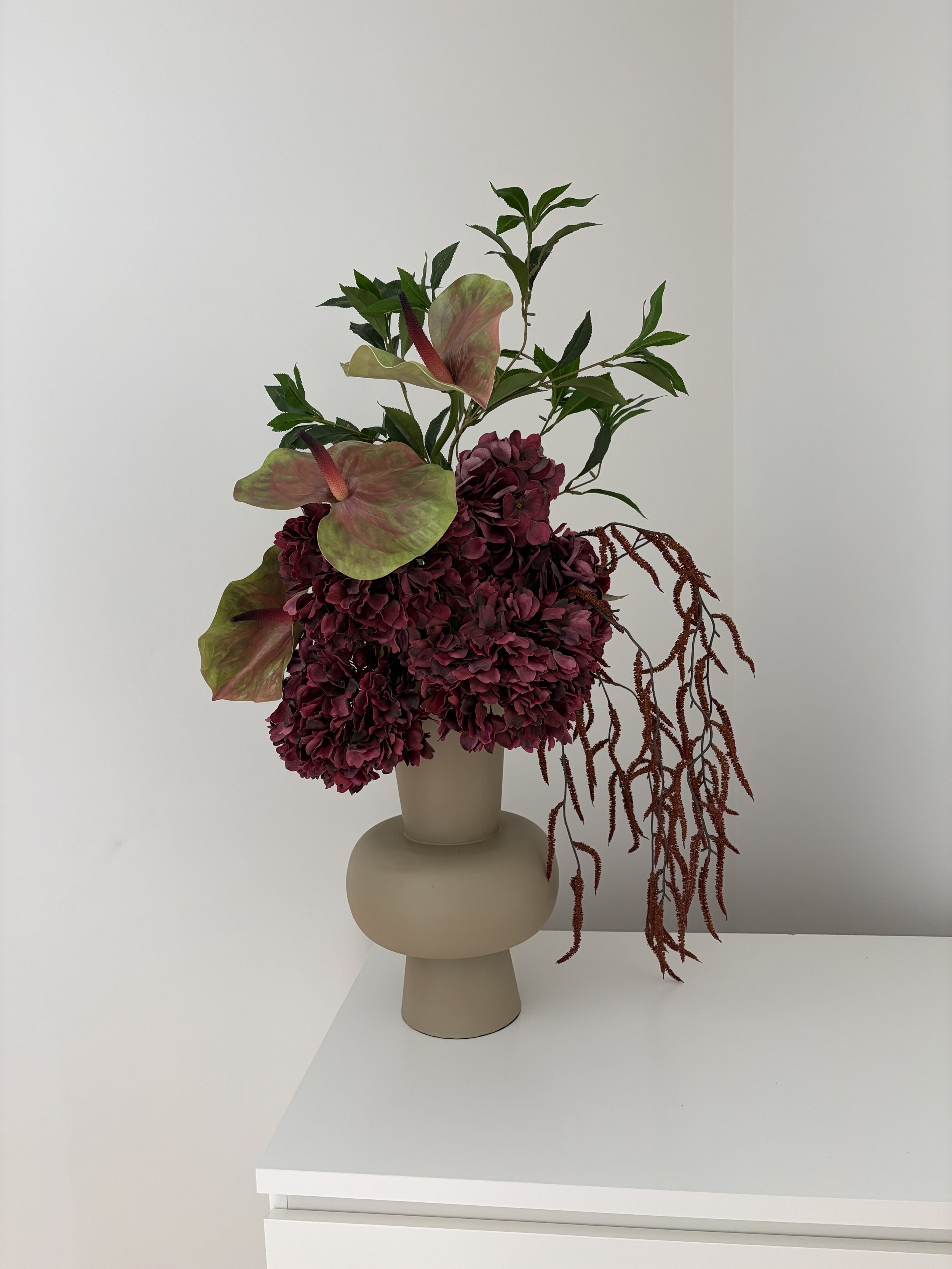 A beige vase with a narrow neck holds a floral arrangement with dark red hydrangeas, green leaves, anthurium with pink and green leaves, and red hanging amaranthus, placed on a white surface against a plain white background.
