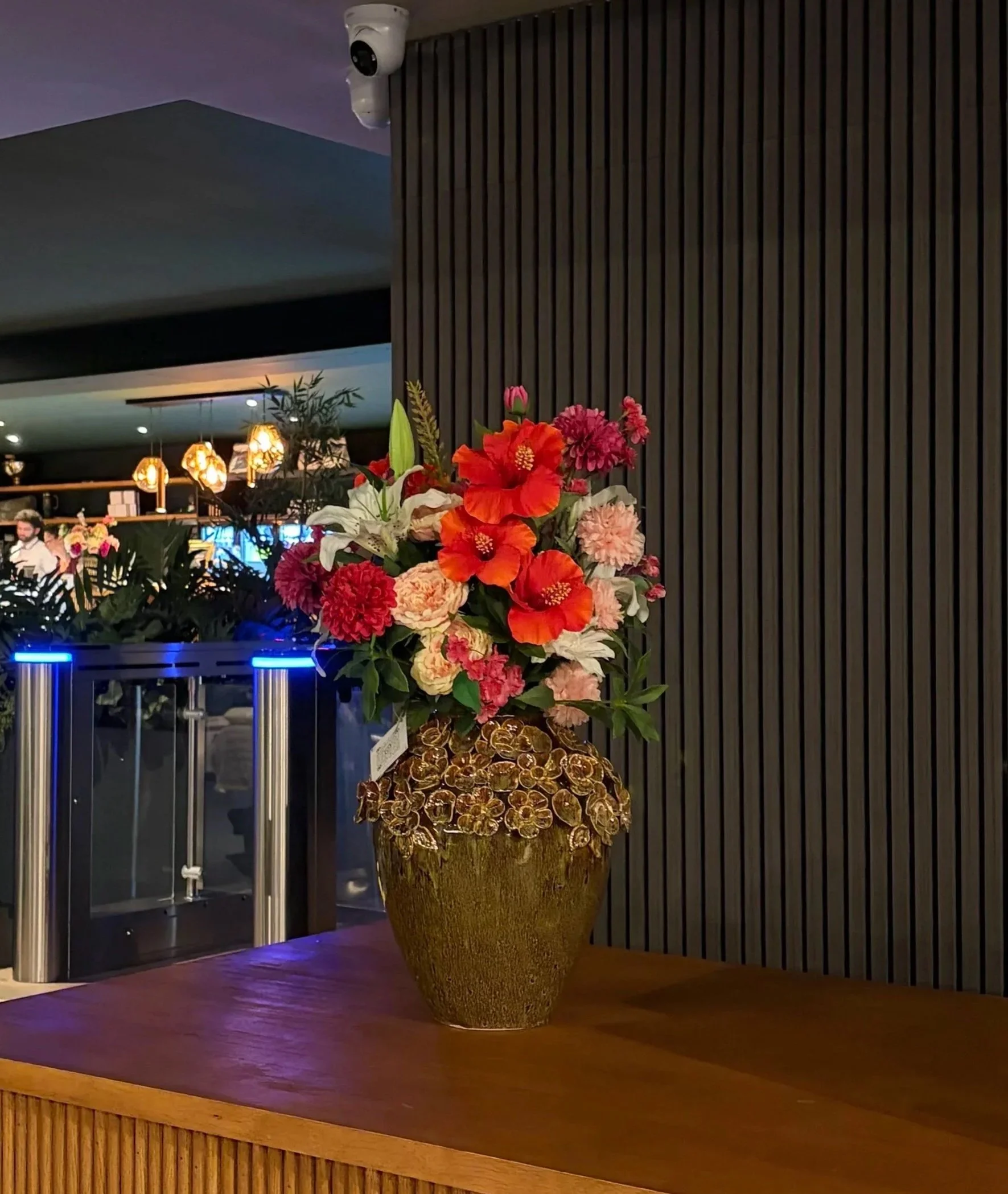 A large decorative flower vase filled with colorful artificial flowers including orange, pink, and white blooms, placed on a wooden stage with a dark background and modern interior decor.