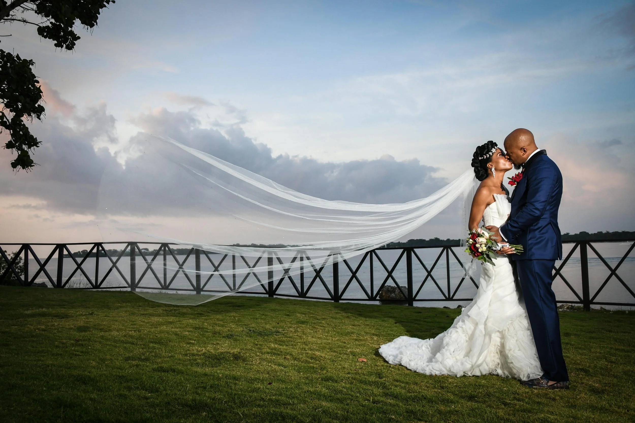 A bride and groom sharing a kiss outdoors by a lake during sunset. The bride is wearing a strapless white wedding gown with a long train and a veil flowing in the wind. The groom is dressed in a dark blue suit with a white shirt and black tie, holding a bouquet of flowers. There is a wooden fence and green grass in the foreground, and clouds and sky with some pink hues in the background.