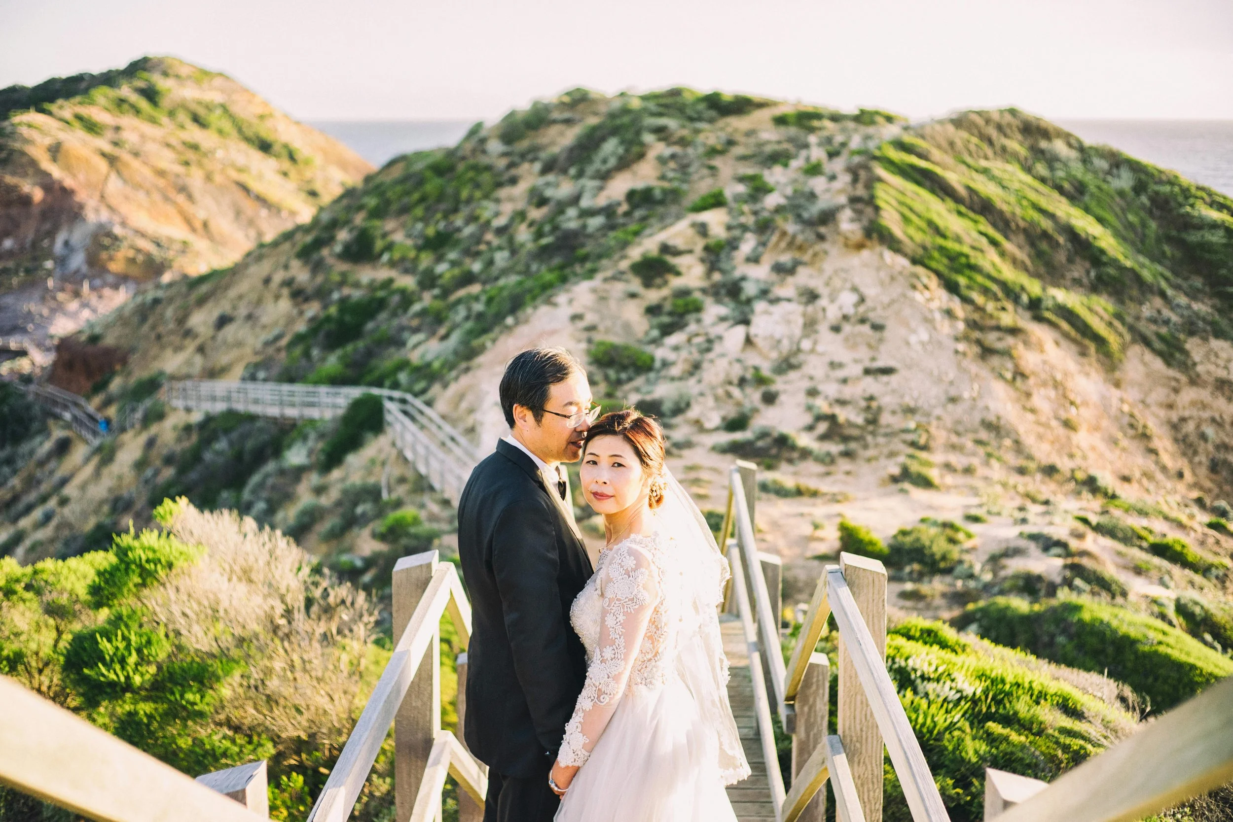 A bride and groom standing on a wooden walkway at the beach with cliffs in the background during sunset.