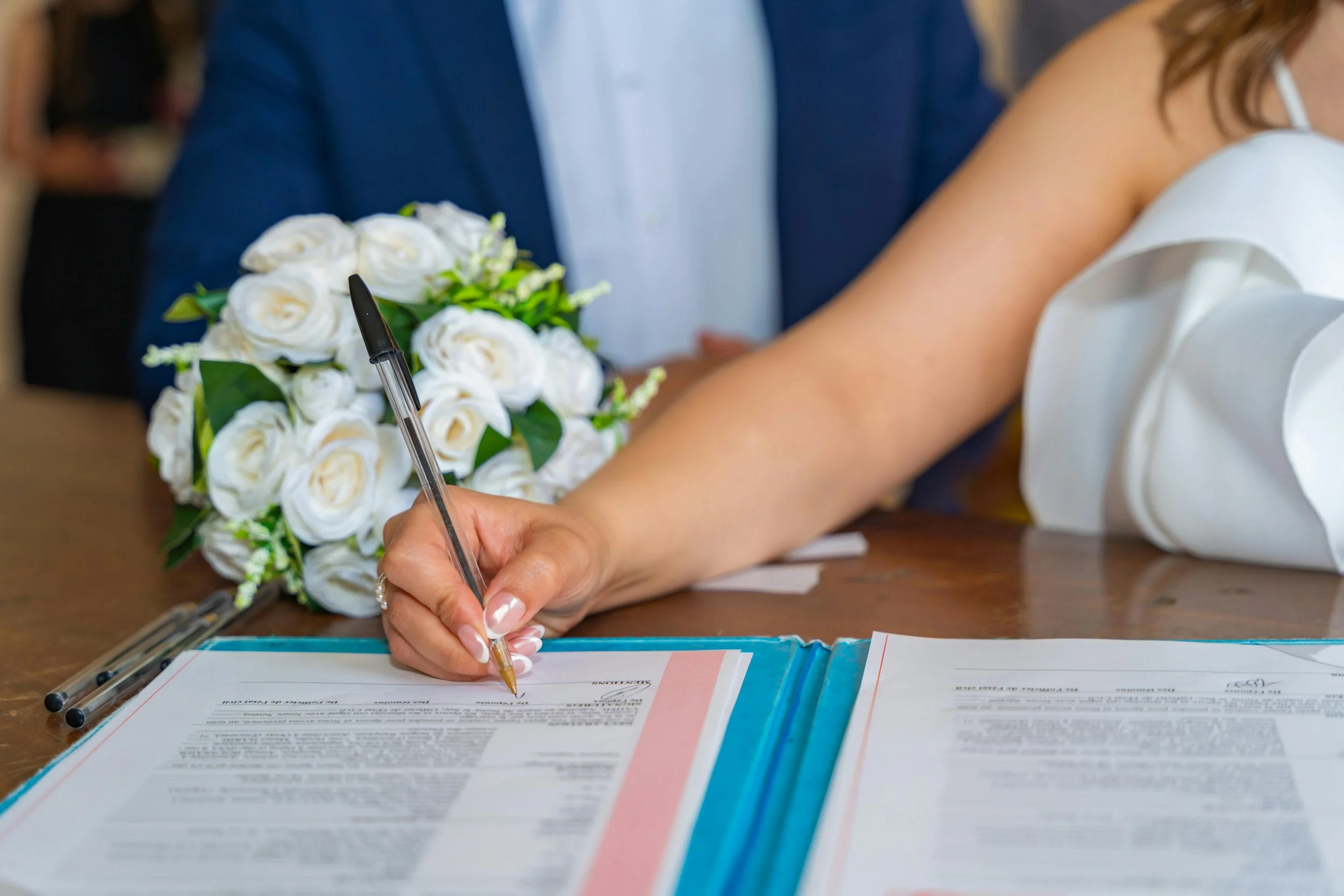 Person signing a document during a formal ceremony, with a bouquet of white roses and two pens on the table.