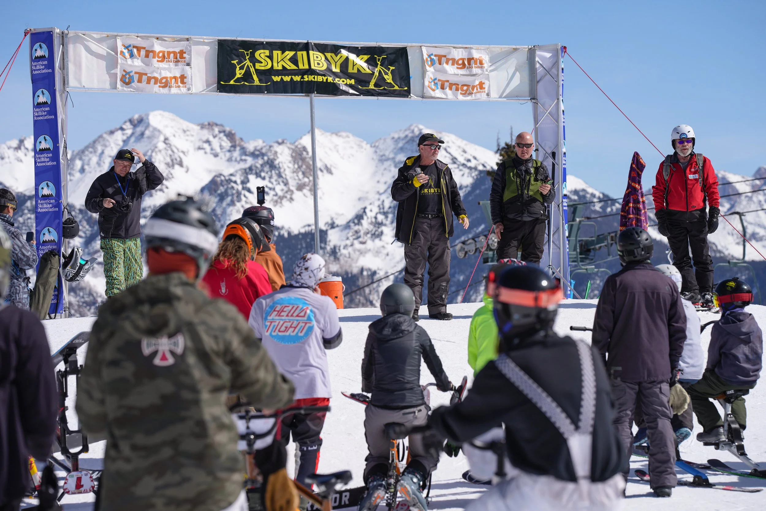 People gathered on a snowy mountain at a ski event, with snow-covered peaks in the background, and a banner with logos and the word 'SKIBYK' at a ski resort.