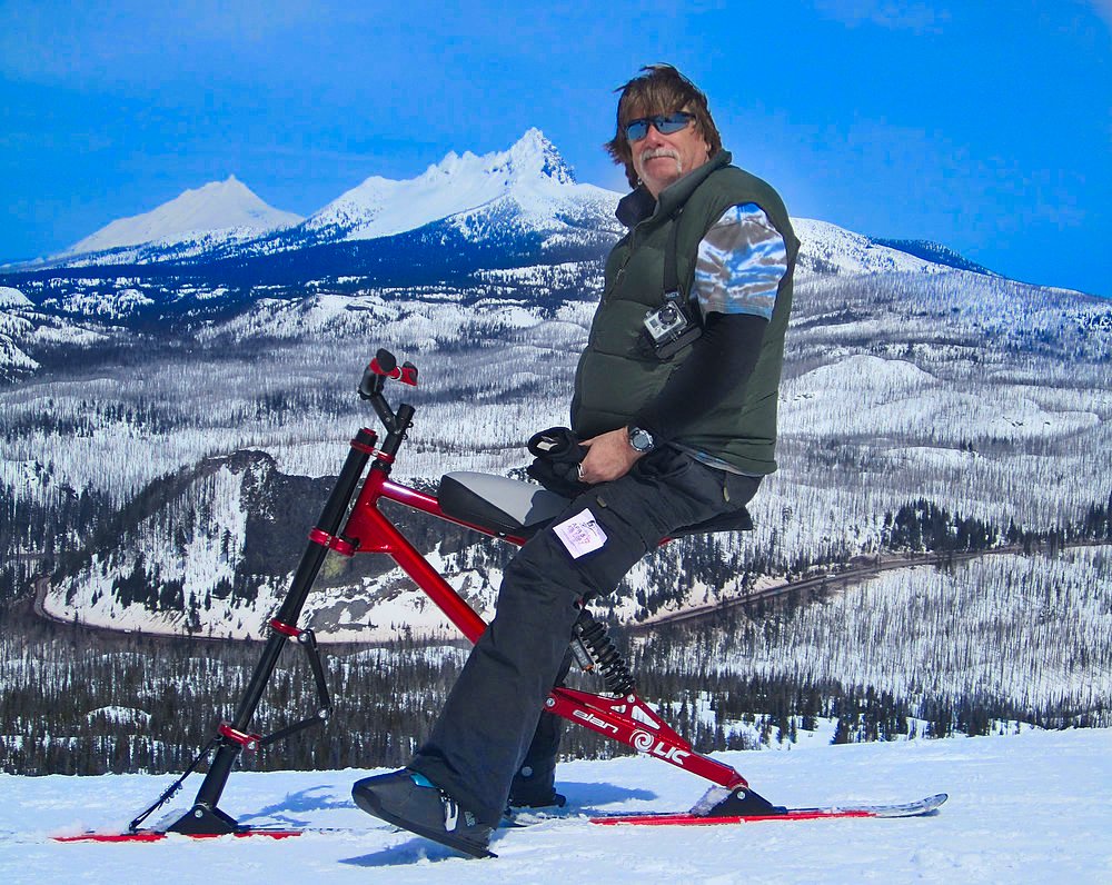 A man with sunglasses and a green vest sitting on a red snow bike on snow-covered terrain with snow-capped mountains in the background.