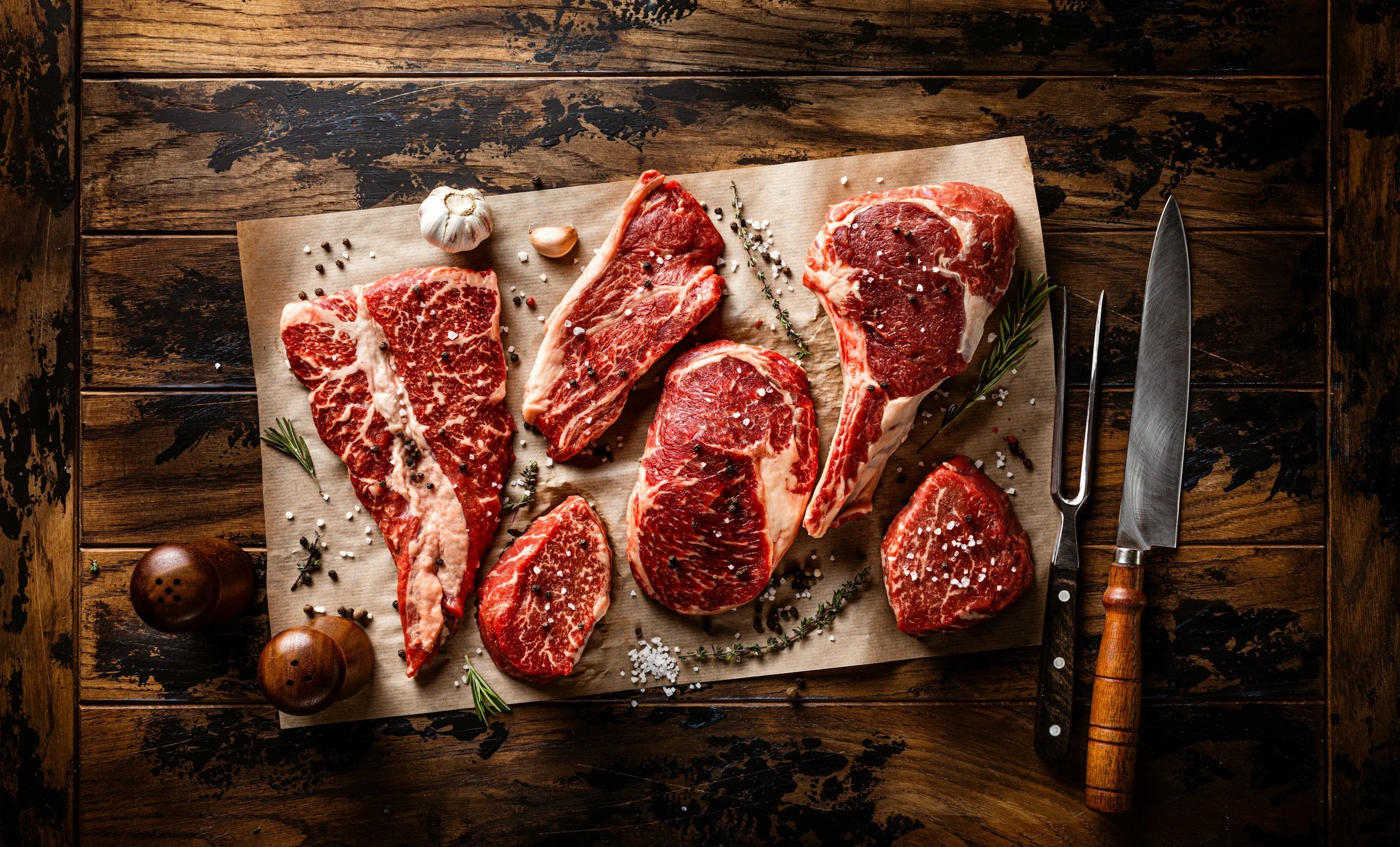 Assorted raw steaks on parchment paper with garlic, herbs, salt, pepper, and a meat fork and knife on a rustic wooden table. Paddock to Plate. Fresh, healthy, grass-fed beef for sale and home-delivery to customer in Brisbane, Queensland.