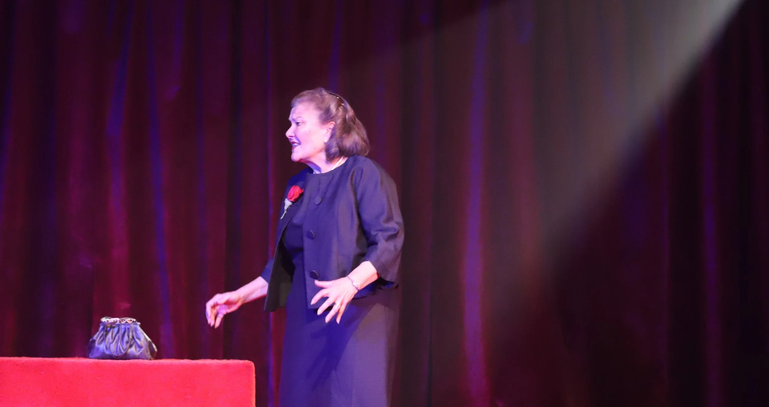 A woman on stage dressed in a black outfit with a red flower pin, standing near a table with a small purse, against a backdrop of dark red curtains, appearing to perform or speak.