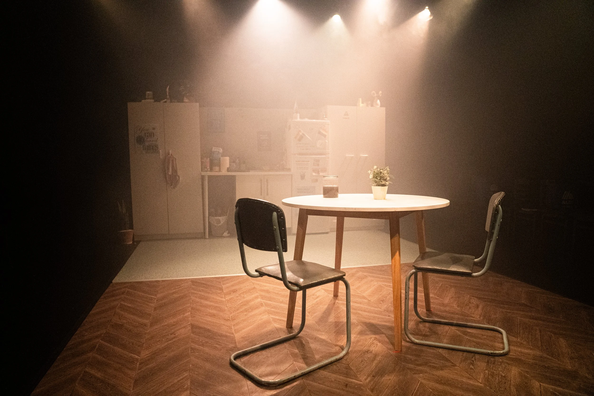 Empty kitchen with a round table and two chairs, a potted plant, and a blurred background of cabinets and kitchen appliances.