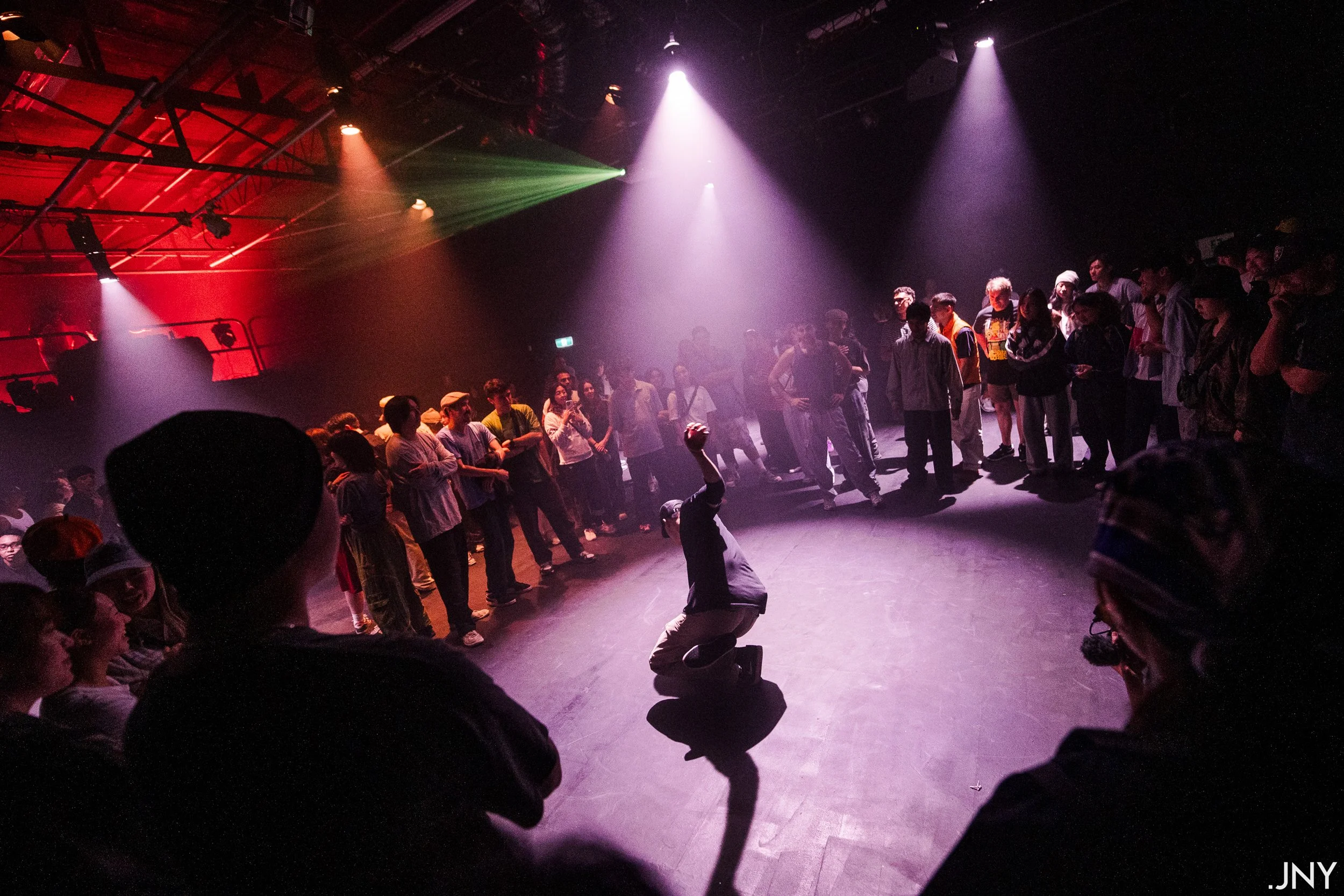 A man performing a dance move in the center of a crowd in a dark, lit dance studio or nightclub with colored stage lights.