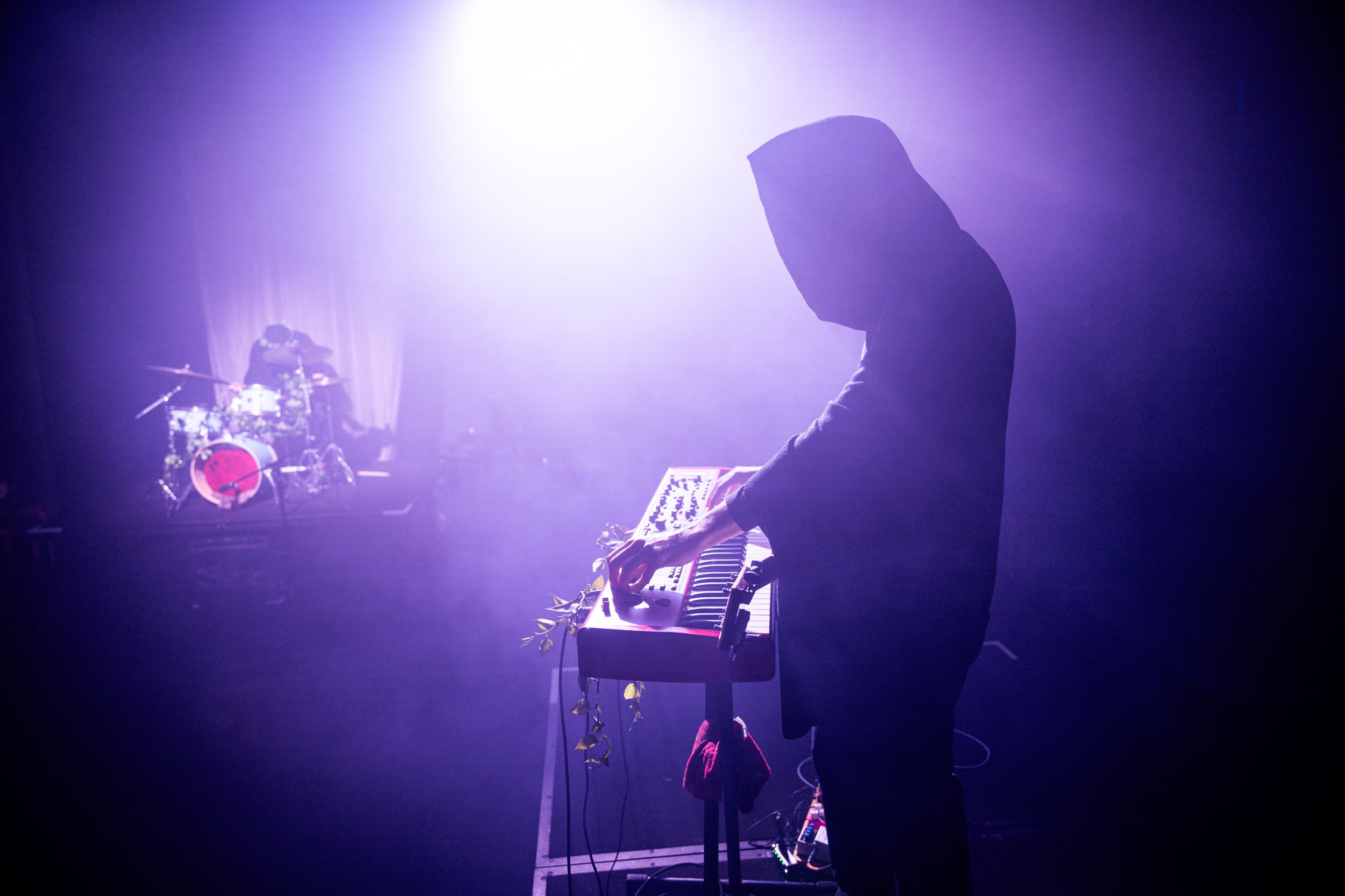 A person wearing a hooded jacket playing a keyboard or synthesizer on stage, with a drum set and a drummer in the background, under dramatic purple stage lighting.