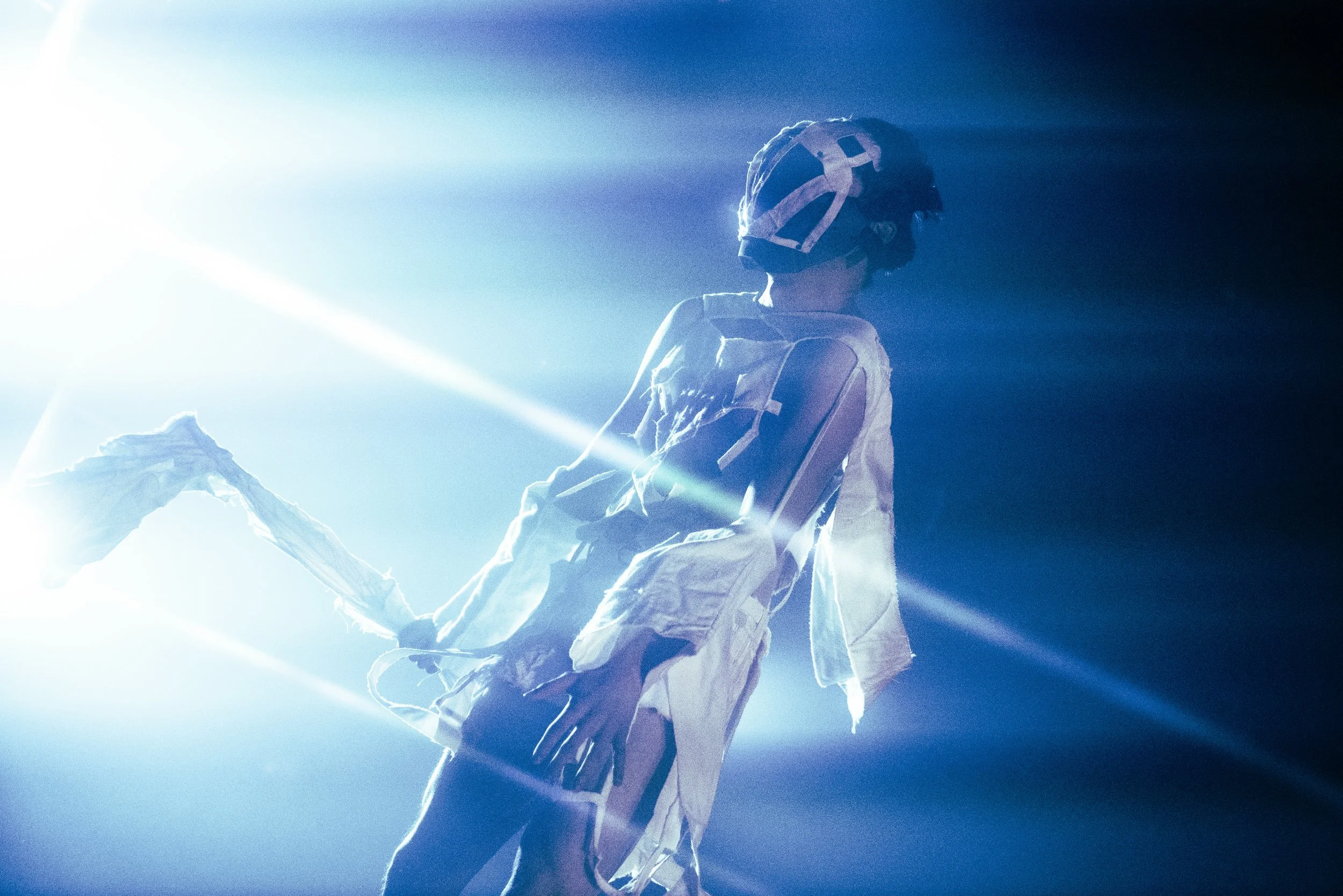 A person in a space suit, sitting on a chair with a helmet, illuminated by bright blue and white light.