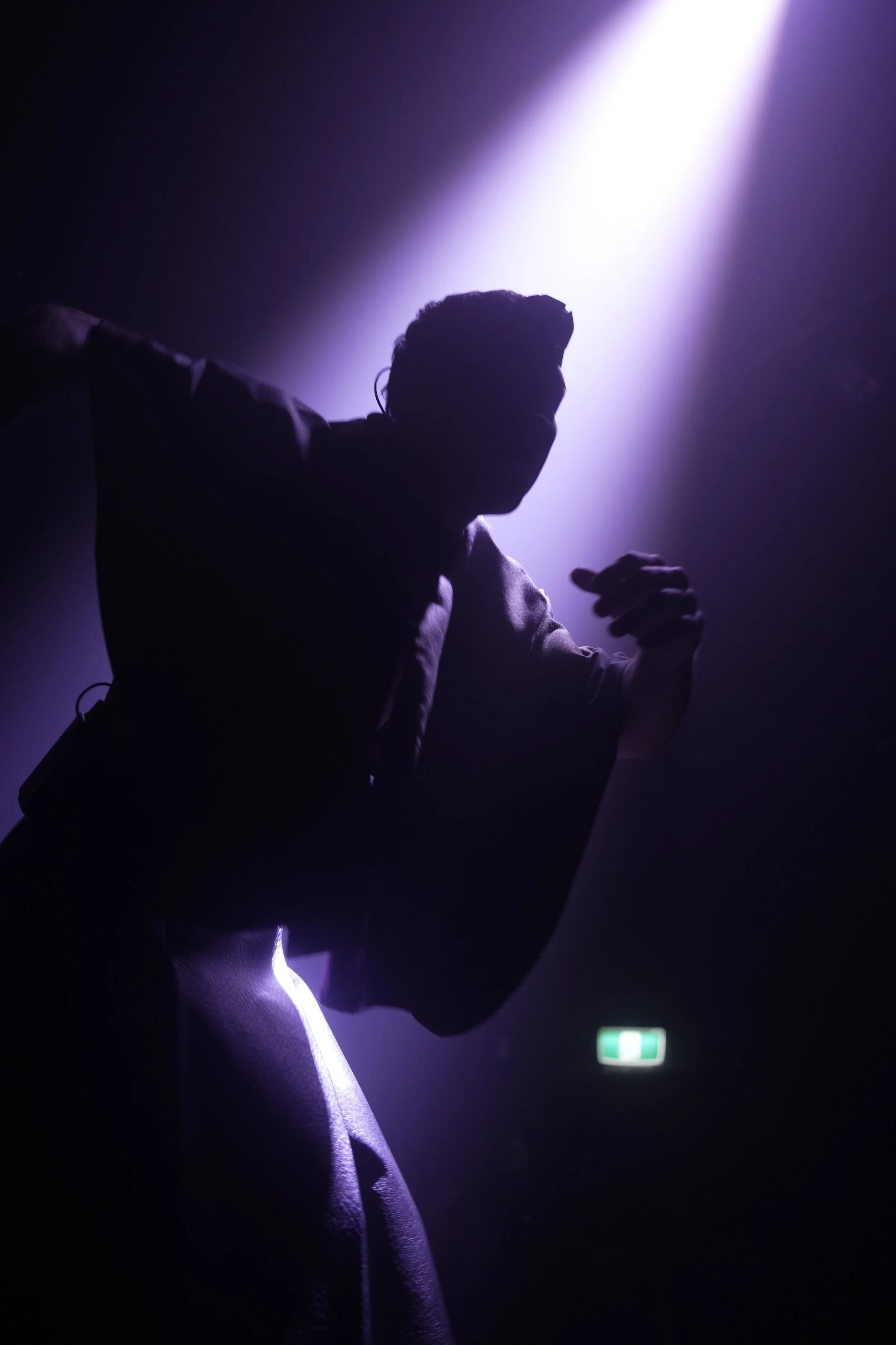 Silhouette of a person dancing or performing on stage with purple lighting and an exit sign in the background.