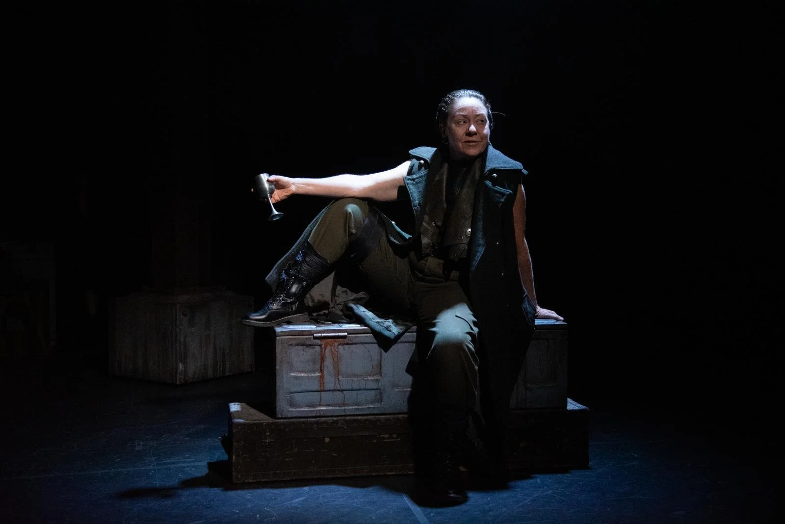 A woman sitting on a wooden chest holding a goblet in her right hand, dressed in military-style clothing and a dark vest, on a dark stage with spotlight