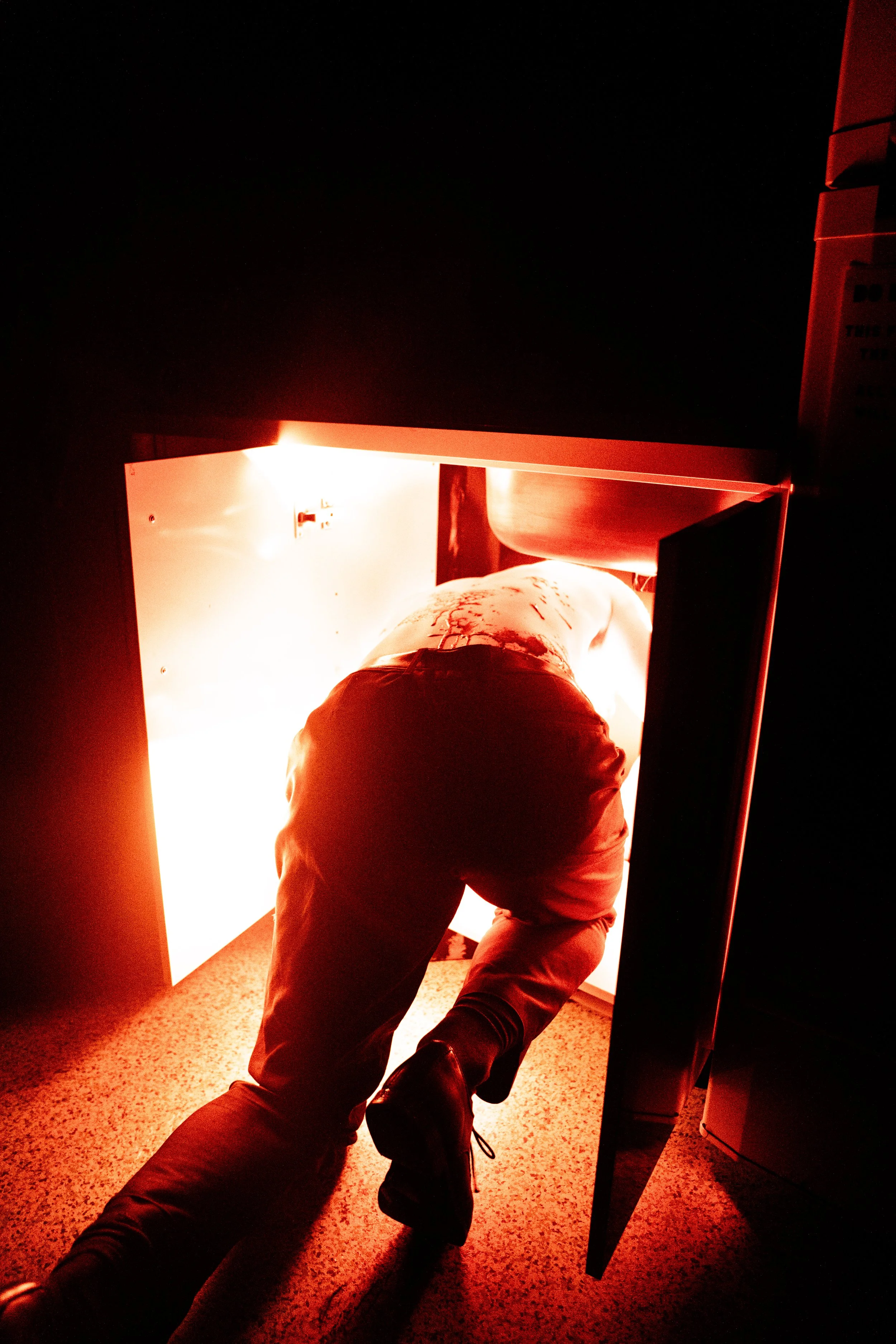 A person crawling into a bright red emergency safety cabinet, with bloodstains on their back and a blood-stained shirt, in a dark environment.