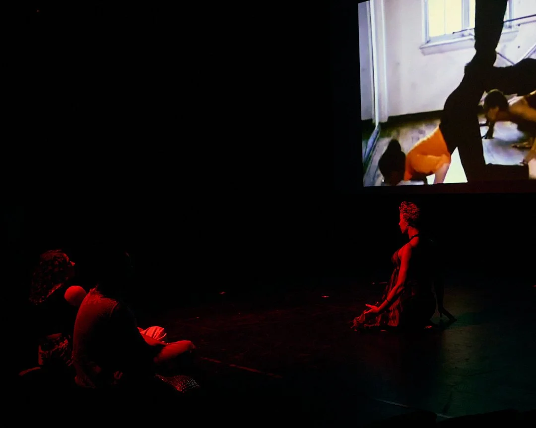 Two performers sitting on the stage facing each other with a large screen displaying a scene in the background.