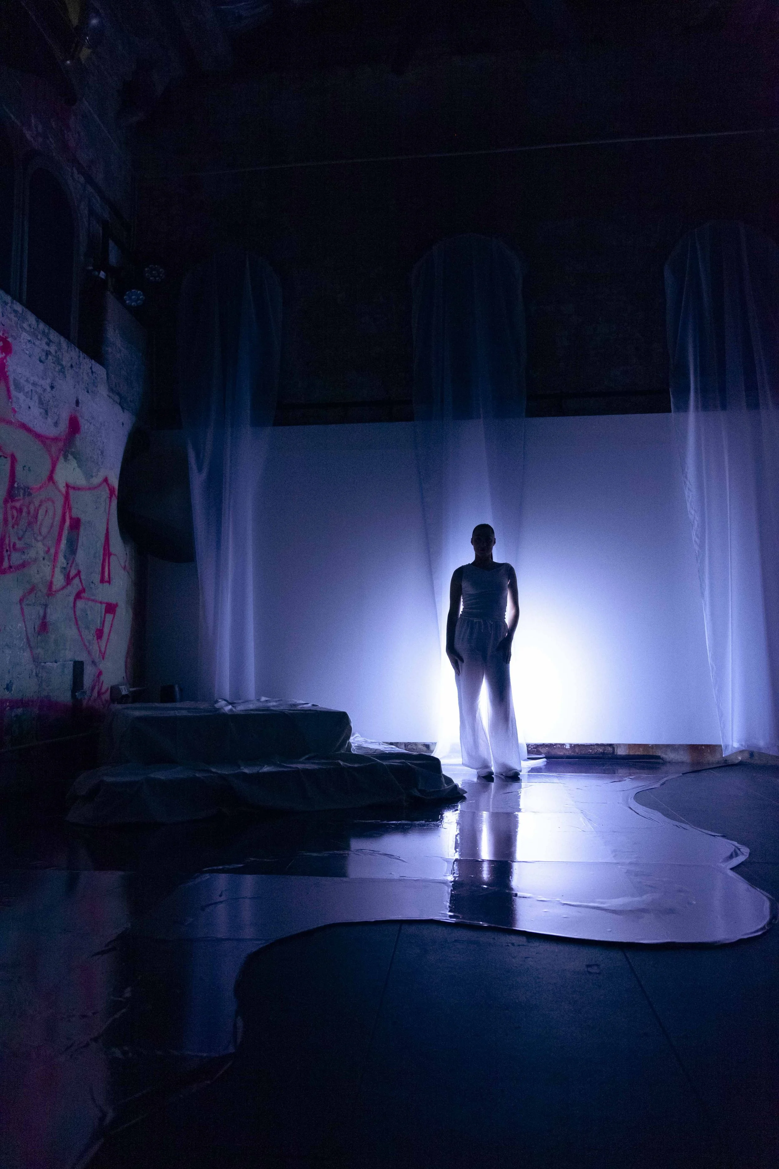 A person standing on a reflective floor in front of a bright light, surrounded by sheer curtains hanging from the ceiling, with graffiti on the wall to the left.