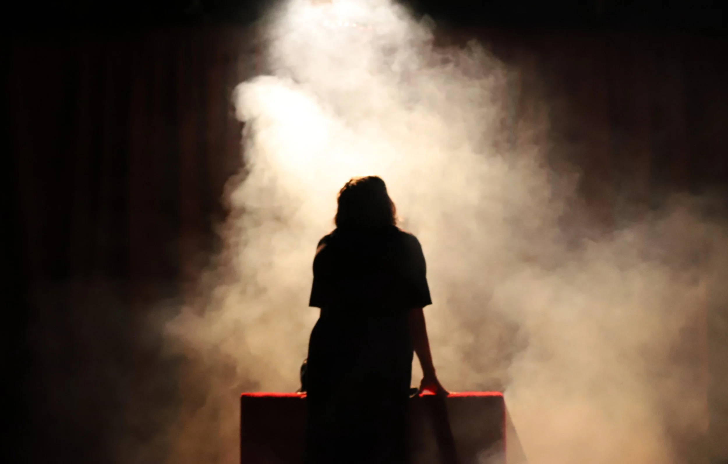 Silhouette of a person sitting on a red bench with smoke and bright light behind them in a dark environment.