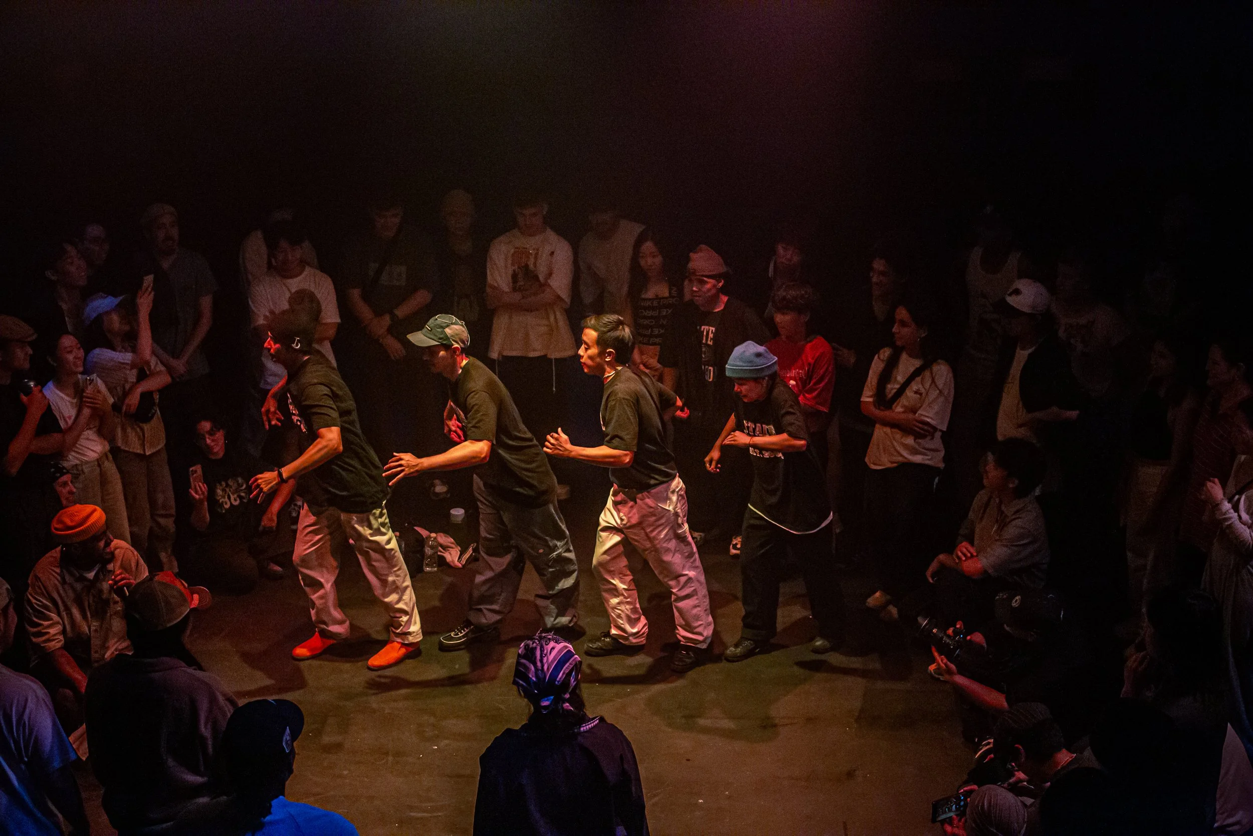 Group of people dancing in a circle on stage, surrounded by spectators watching and filming, dim lighting with focus on the dancers.