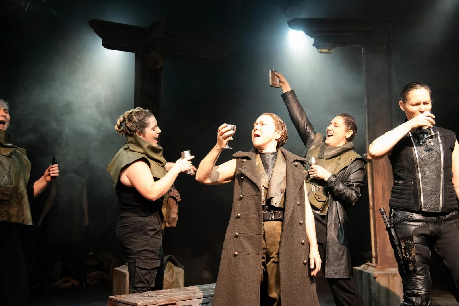 Actors performing on stage, dressed in military-style costumes, holding glasses and mugs, celebrating with smiles.
