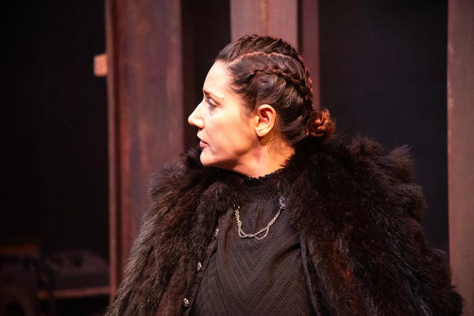 Side profile of a woman with braided hair wearing a black fur coat and necklace, standing indoors.
