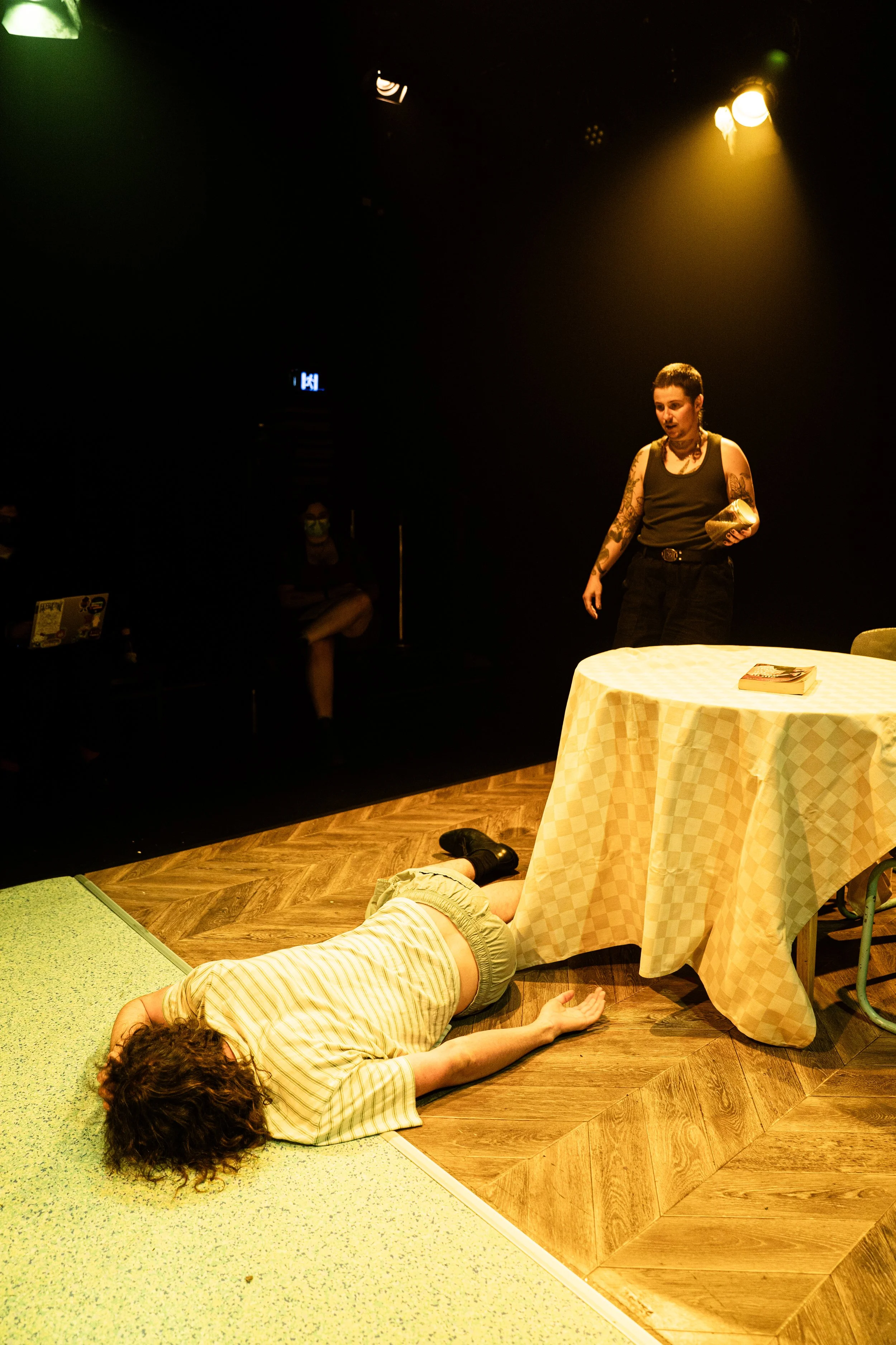 A woman with tattoos standing next to a table, looking down at a person lying on the floor underneath the table, on a stage with a dark background and stage lighting.