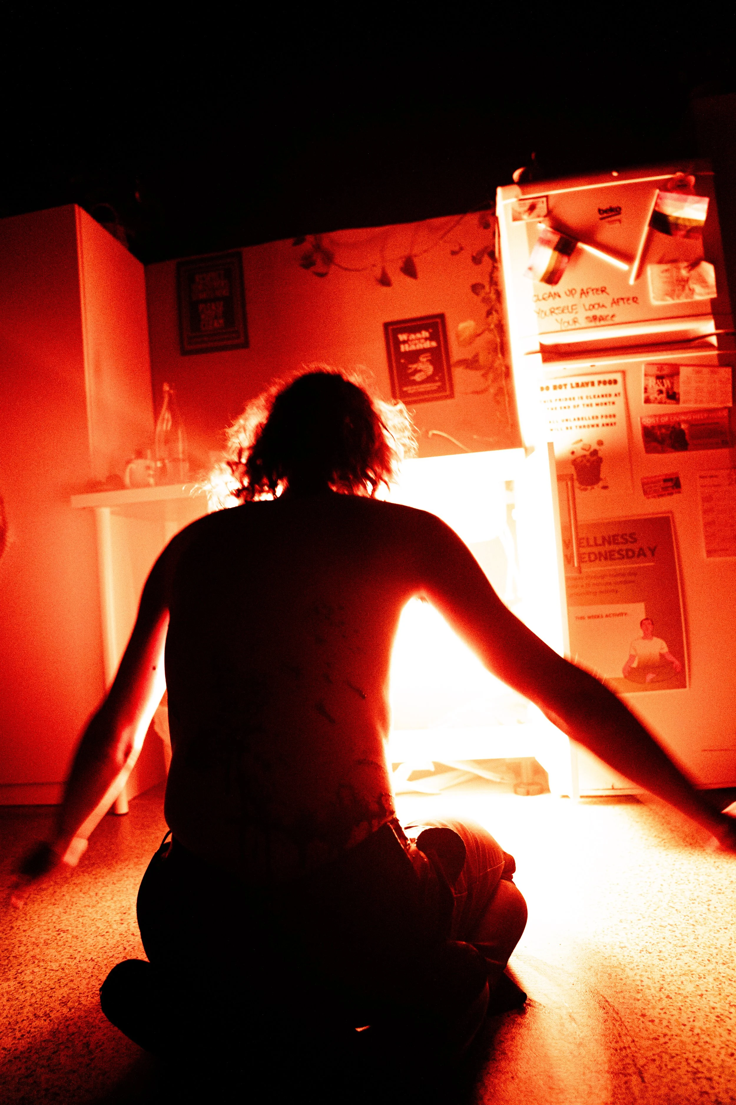 Person with curly hair sitting on the floor in front of a bright red light, with posters and notices on the wall behind.