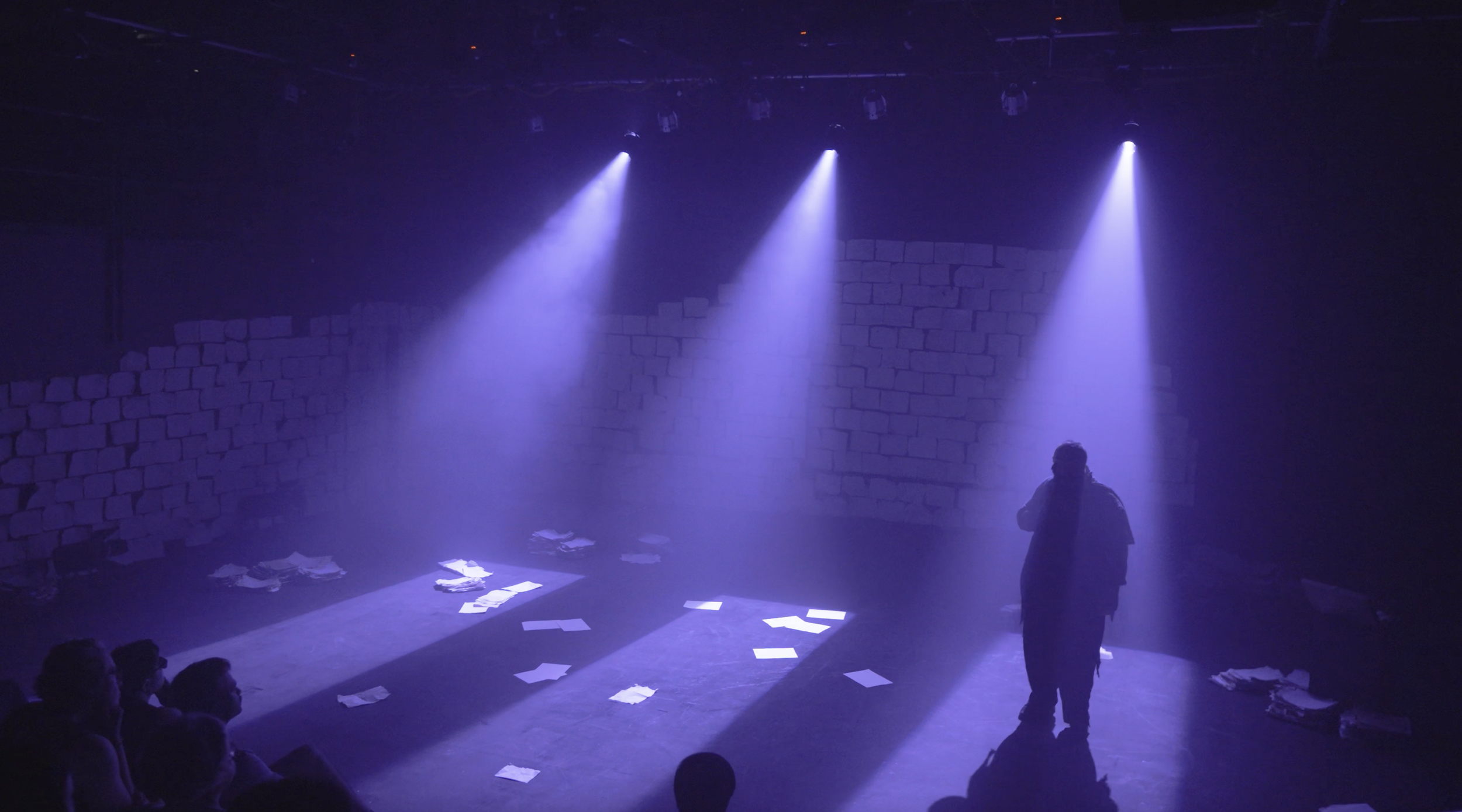 A person stands on stage illuminated by purple spotlights, with scattered papers on the floor and a brick wall in the background. Audience members are visible in the foreground.