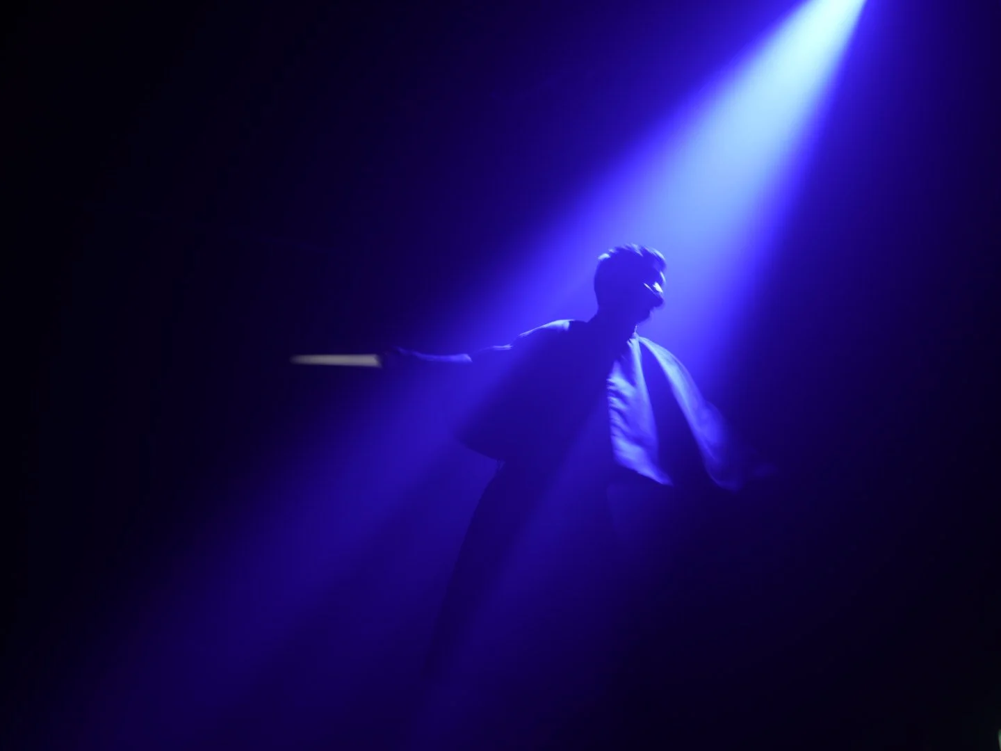 A silhouette of a performer on stage illuminated by blue and purple lighting, with arms outstretched and t-shirt in a dark setting.