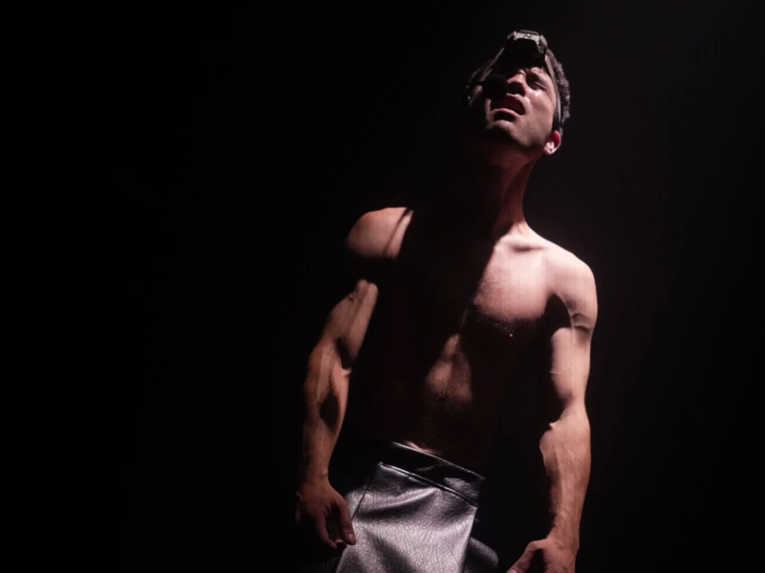 A shirtless man with headphones and a headlamp looking upward, with dramatic lighting creating shadows on his face and upper body.