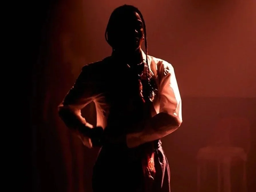 A person with long hair in a ponytail standing on a dimly lit stage, wearing a white shirt and dark pants, facing slightly to the right with arms crossed.