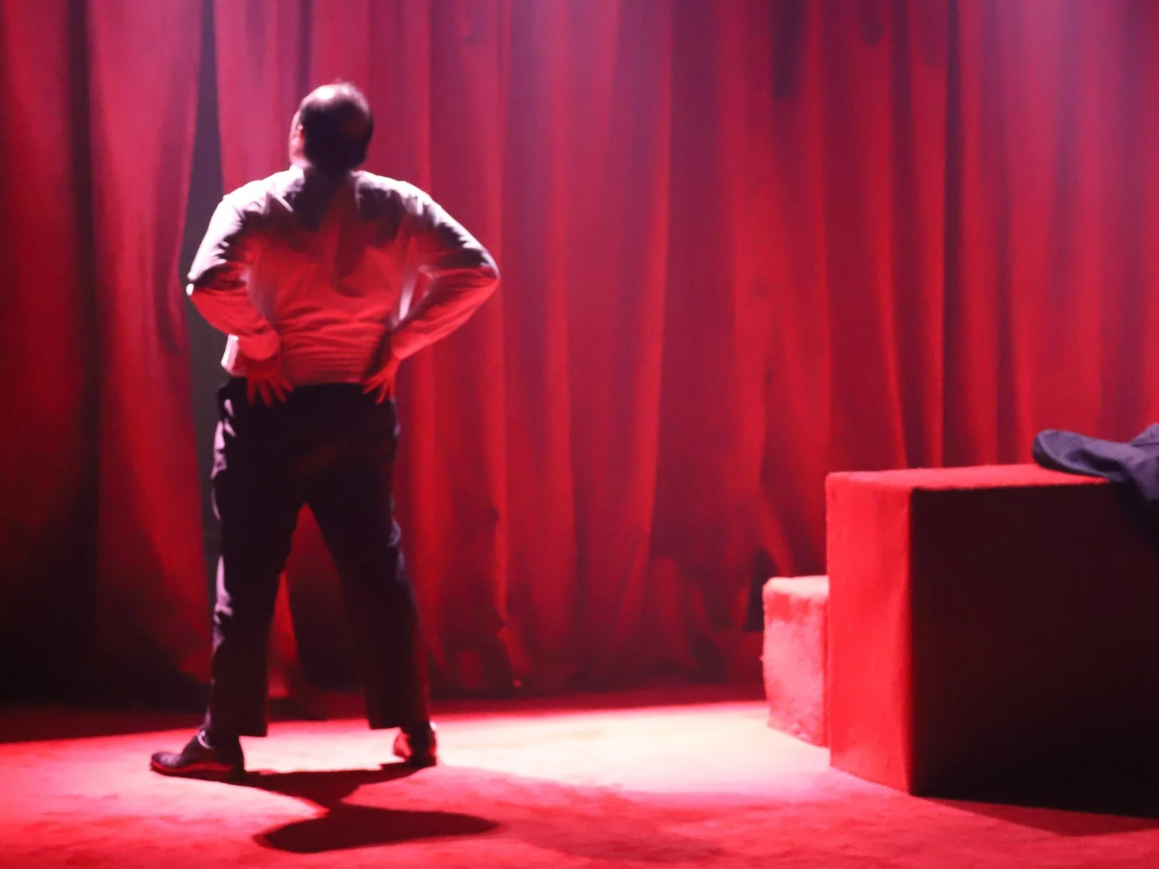 A person with short hair standing on a stage with red curtains in the background, facing away from the camera, with hands on hips, illuminated by stage lighting.