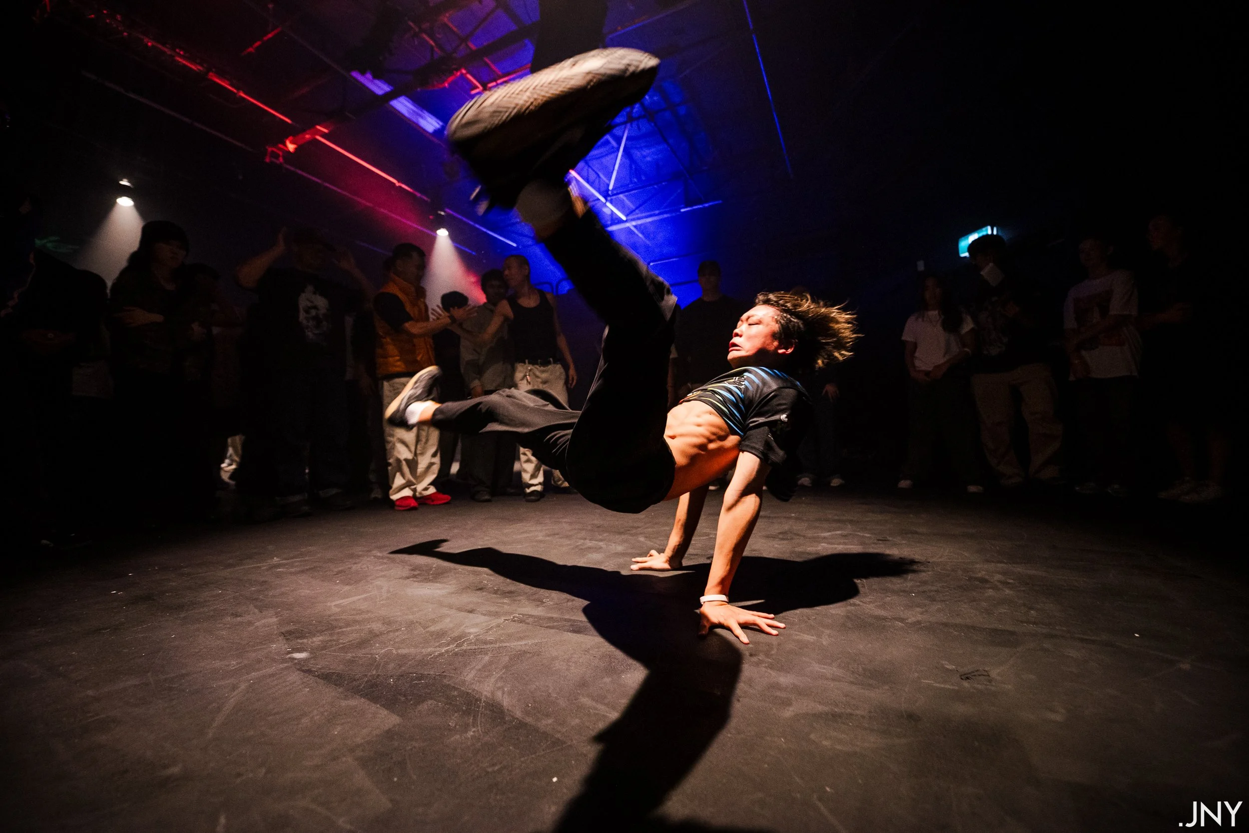 A male breakdancer performing a move on a dark stage with colorful lighting, surrounded by an audience of spectators in casual clothing.