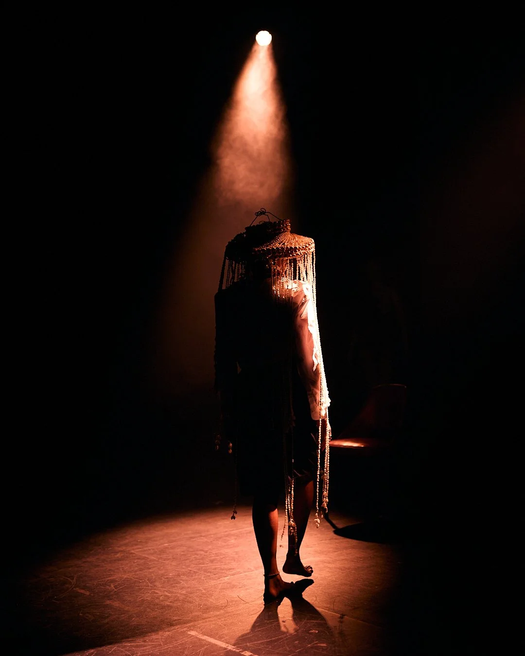 Performer standing on stage under a spotlight, wearing a costume with long hanging beads or strands, in a dark setting.