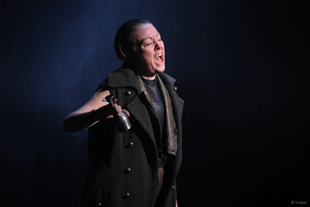 A woman with short dark hair is holding a gun and shouting or screaming, wearing a dark vest and jacket against a dark background.