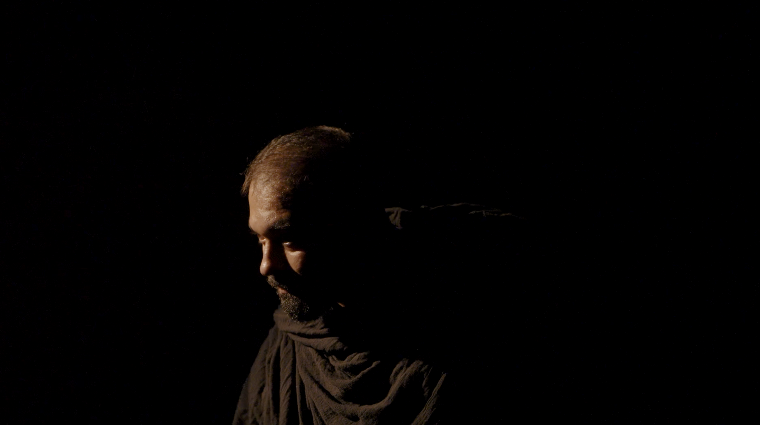A man with short hair and a beard is partially illuminated, with his face turned to the left, in a dark setting with minimal lighting highlighting his profile.