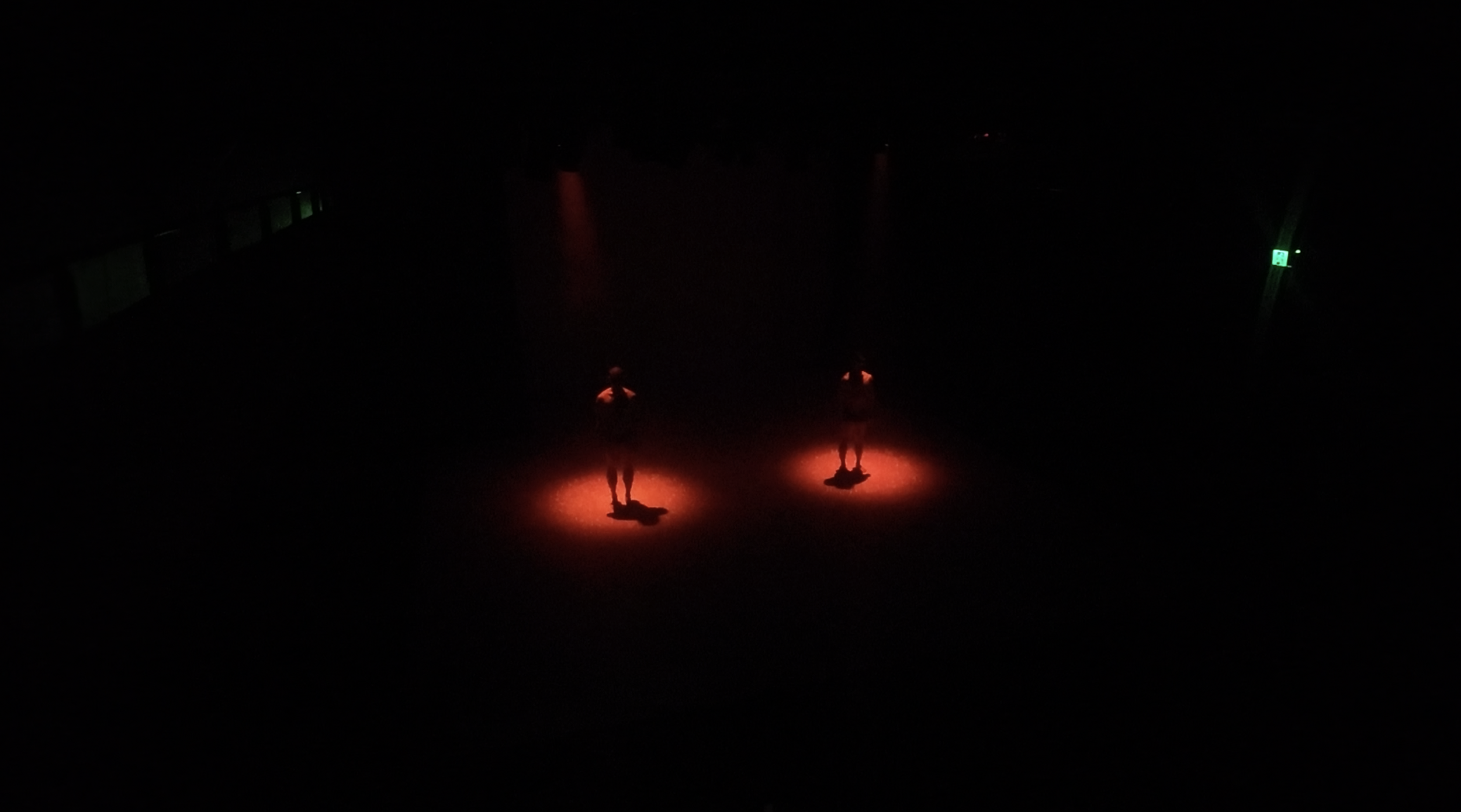 Two people stand illuminated by red lights on the floor in a dark room, with a green emergency exit sign visible on the right side.