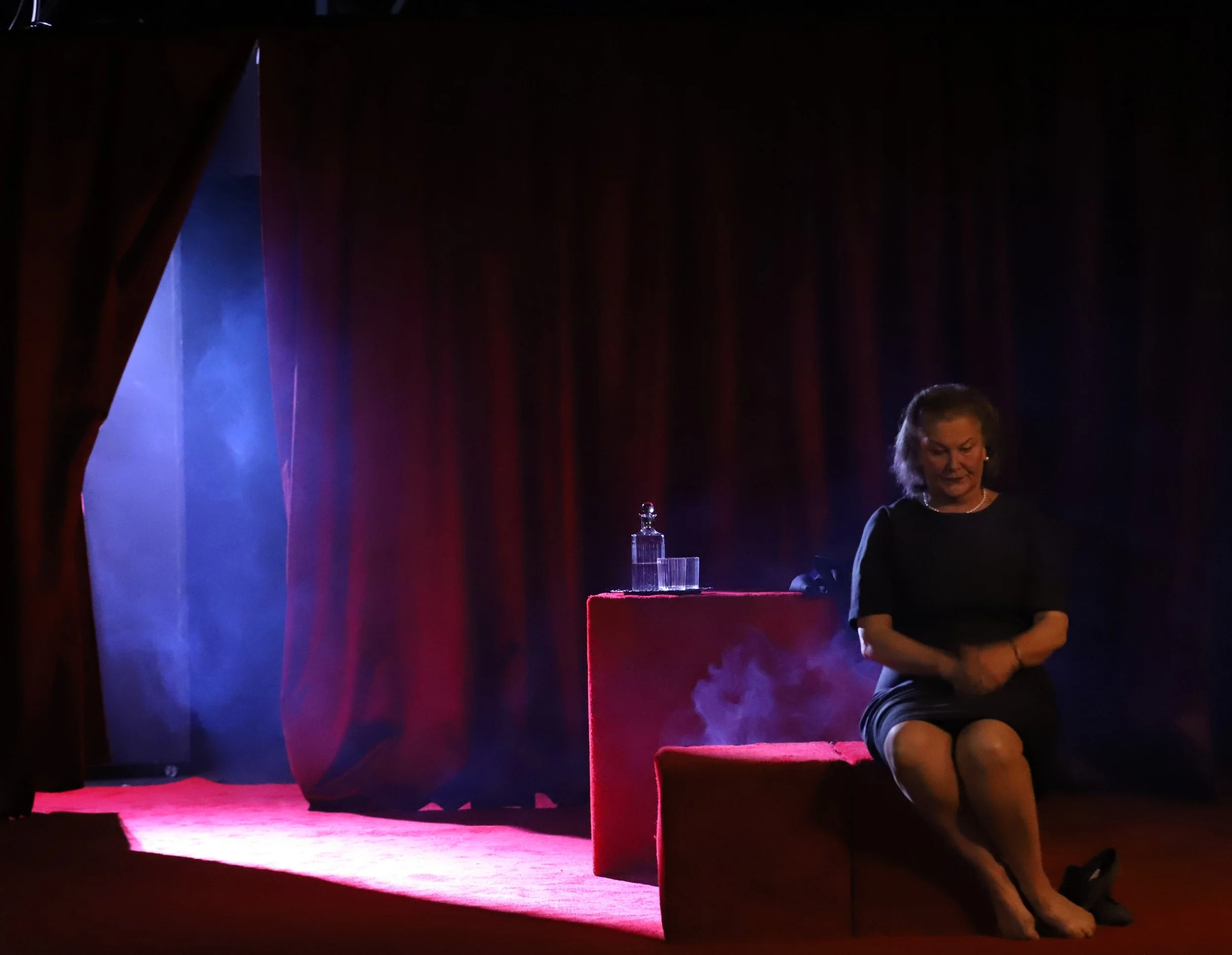 A woman sitting on a red stage box with a small table next to her, on which there is a bottle, a glass, and a decanter, with red curtains in the background and stage lighting creating a moody atmosphere.