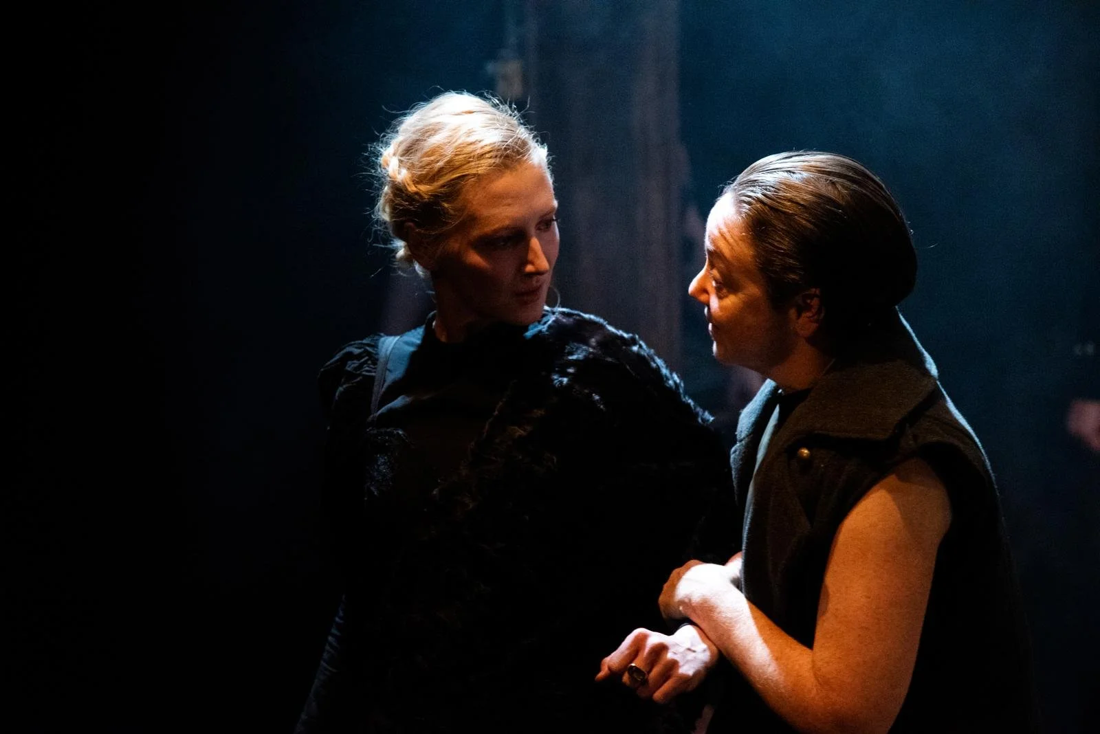 Two women in dark clothing are facing each other and holding hands, one is looking at the other, with a dark background.