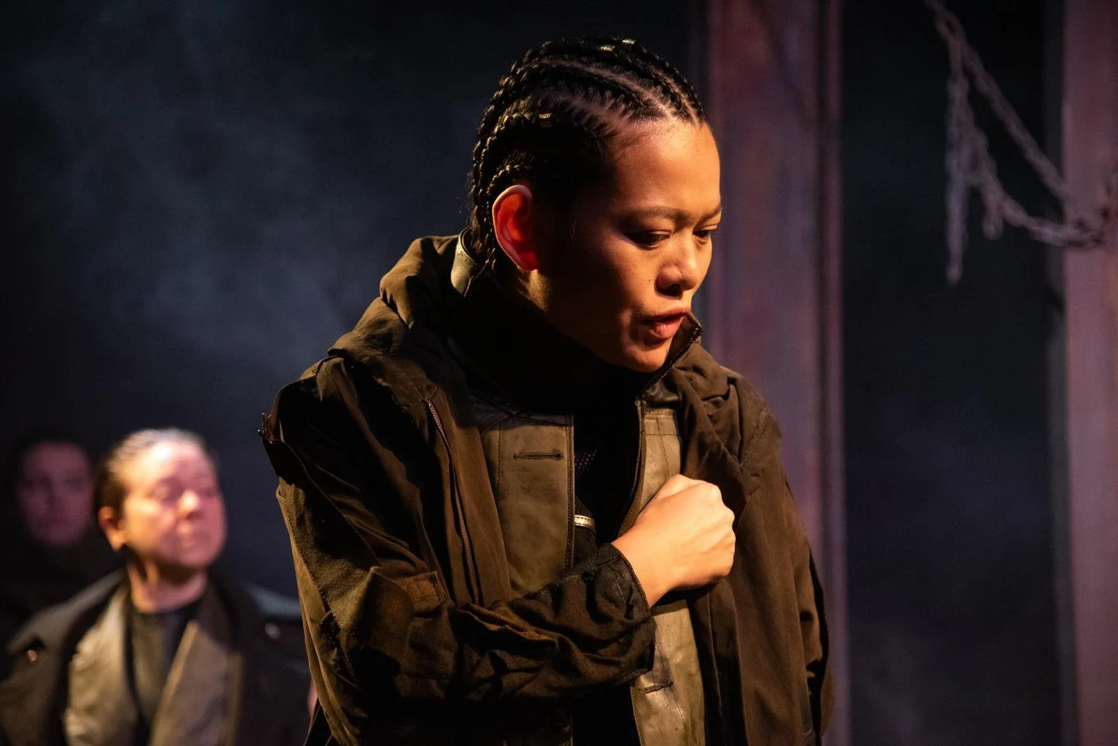 A woman with braided hair looks down and appears emotional, with her hand on her chest. In the background, a man with glasses and a woman with blonde hair look on. The scene is dark with moody lighting, suggesting a dramatic moment.