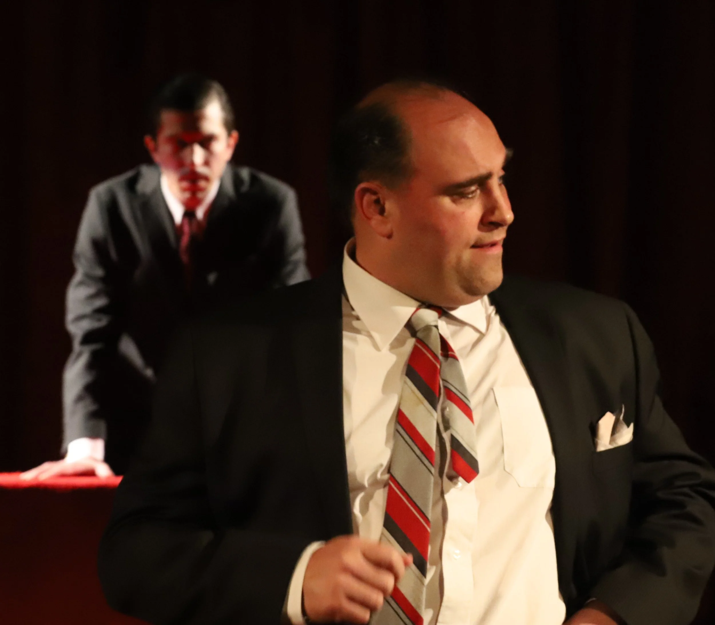 Scene from a play or film with two men in suits, one in the foreground with a confused or displeased expression, and a younger man in the background leaning on a desk, both in formal attire.