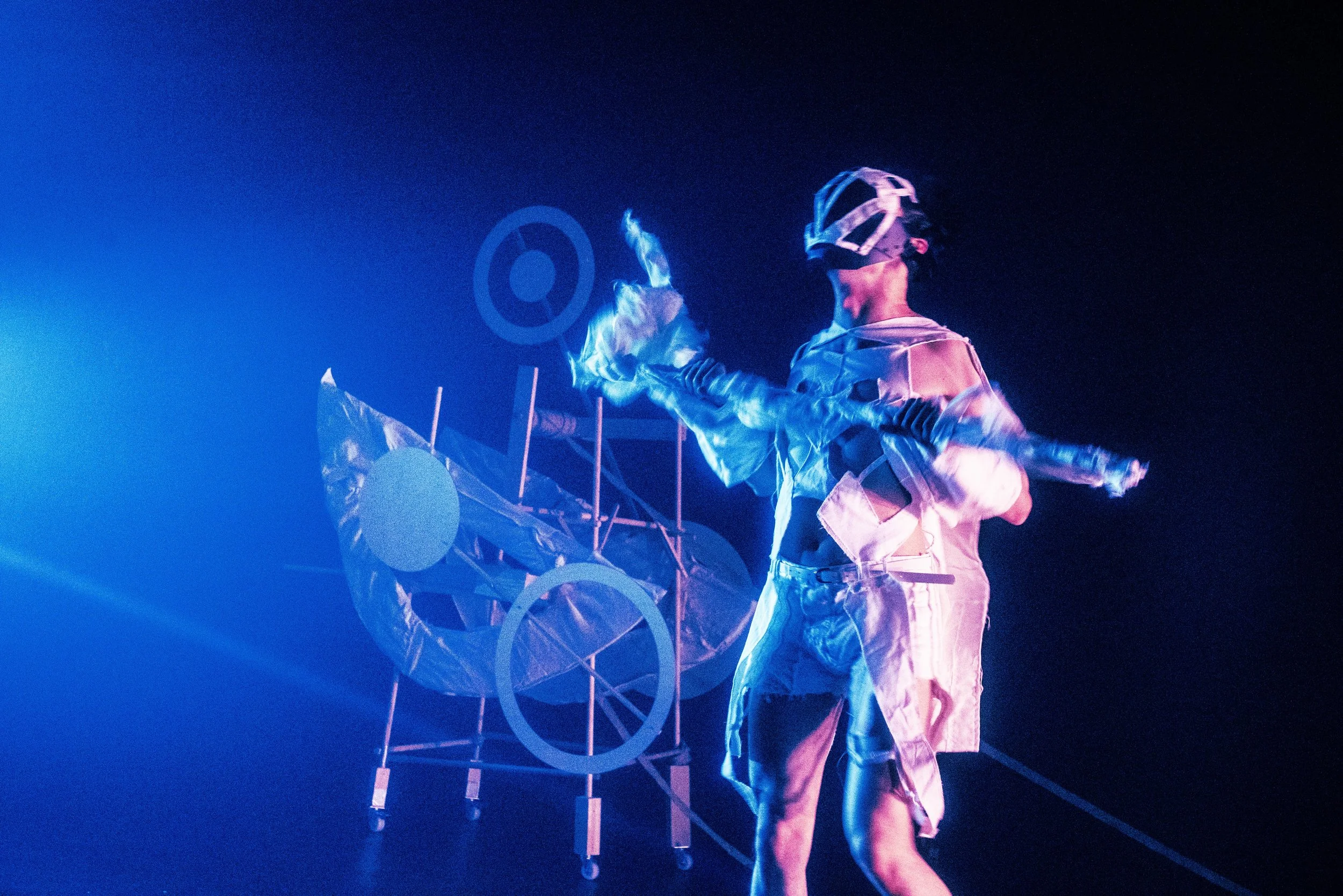 Performer on stage with a futuristic outfit and a visor, manipulating glowing light in front of a complex structure under blue and purple lighting.