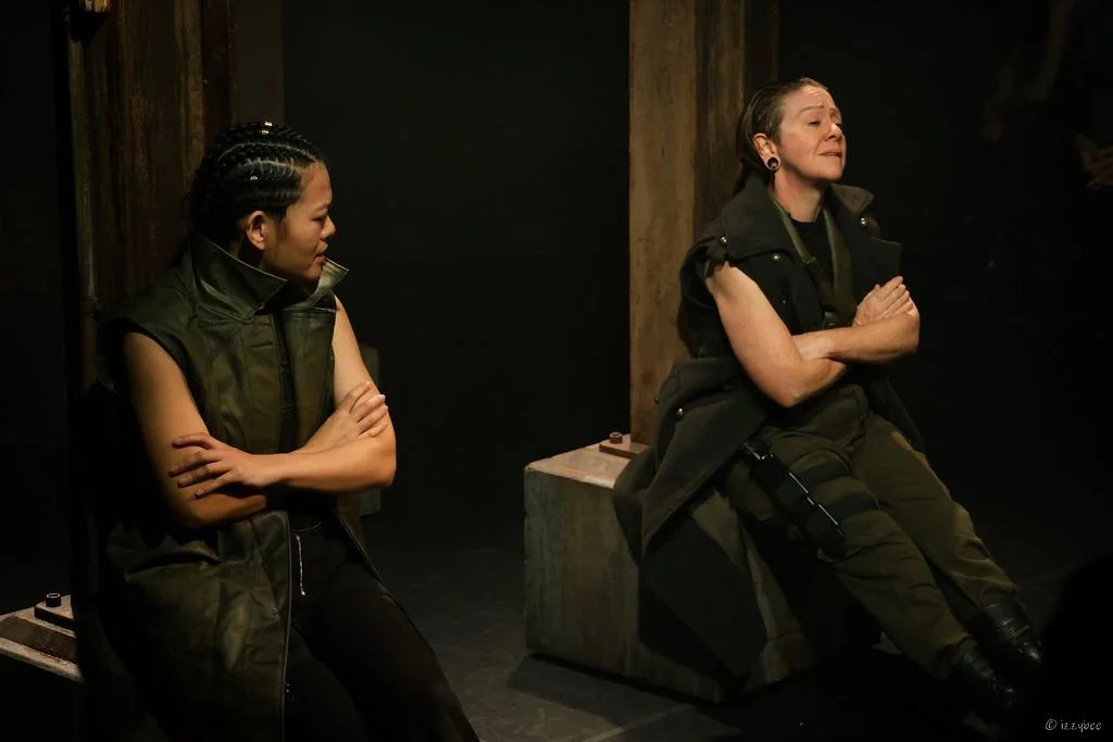 Two women dressed in military-style clothing sitting with arms crossed in a dark room, appearing to be in a moment of contemplation or emotion.
