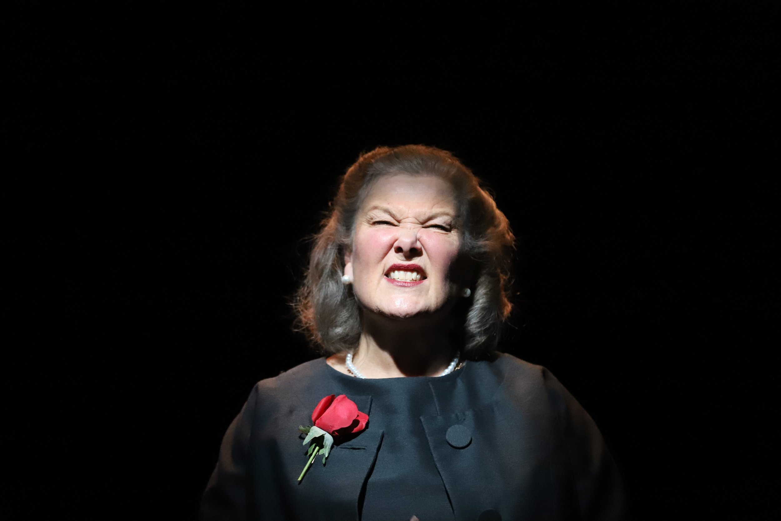 An older woman with gray hair, wearing a dark outfit with a pearl necklace and earrings, grimacing with her eyes squinted and lips curled, standing against a black background with a red rose pinned to her dress.