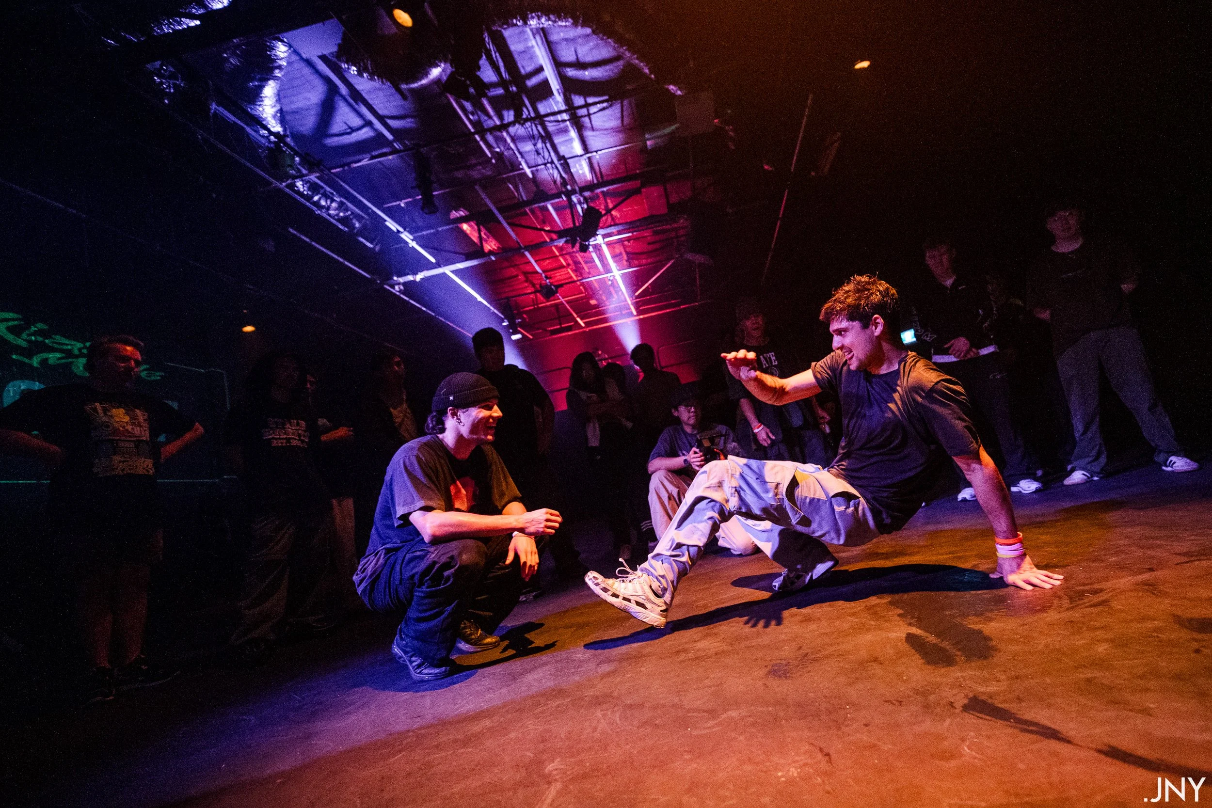 Two young men breakdancing on a stage illuminated with colorful lights, while a group of people watch in the background.